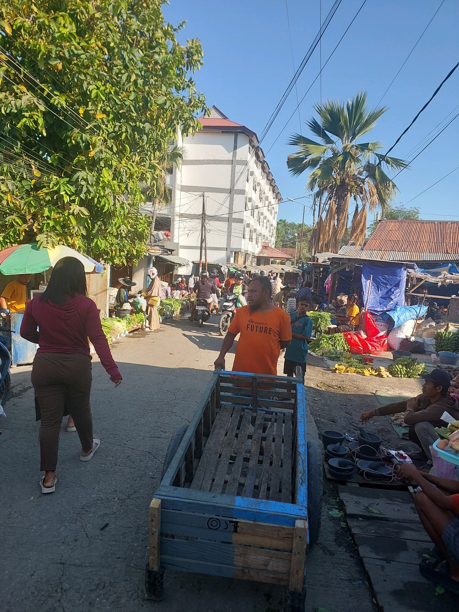 The image depicts a bustling outdoor market scene, likely in Kupang, Indonesia, characterized by its humid tropical climate. In the foreground, a man in an orange t-shirt with the words "POSITIVE FUTURE" is pushing a wooden cart. To his left, a woman in a maroon hoodie and brown pants walks away from the camera. The market is filled with various stalls selling fresh produce, including bananas and leafy green vegetables. Several individuals are seen browsing and shopping. In the background, a multi-story building with white walls and a red roof stands prominently, suggesting an urban setting. A large palm tree, indicative of the tropical environment, is visible behind the market stalls. Power lines crisscross the sky, typical of urban infrastructure. The overall atmosphere is lively and dynamic, portraying a typical day at a local market.