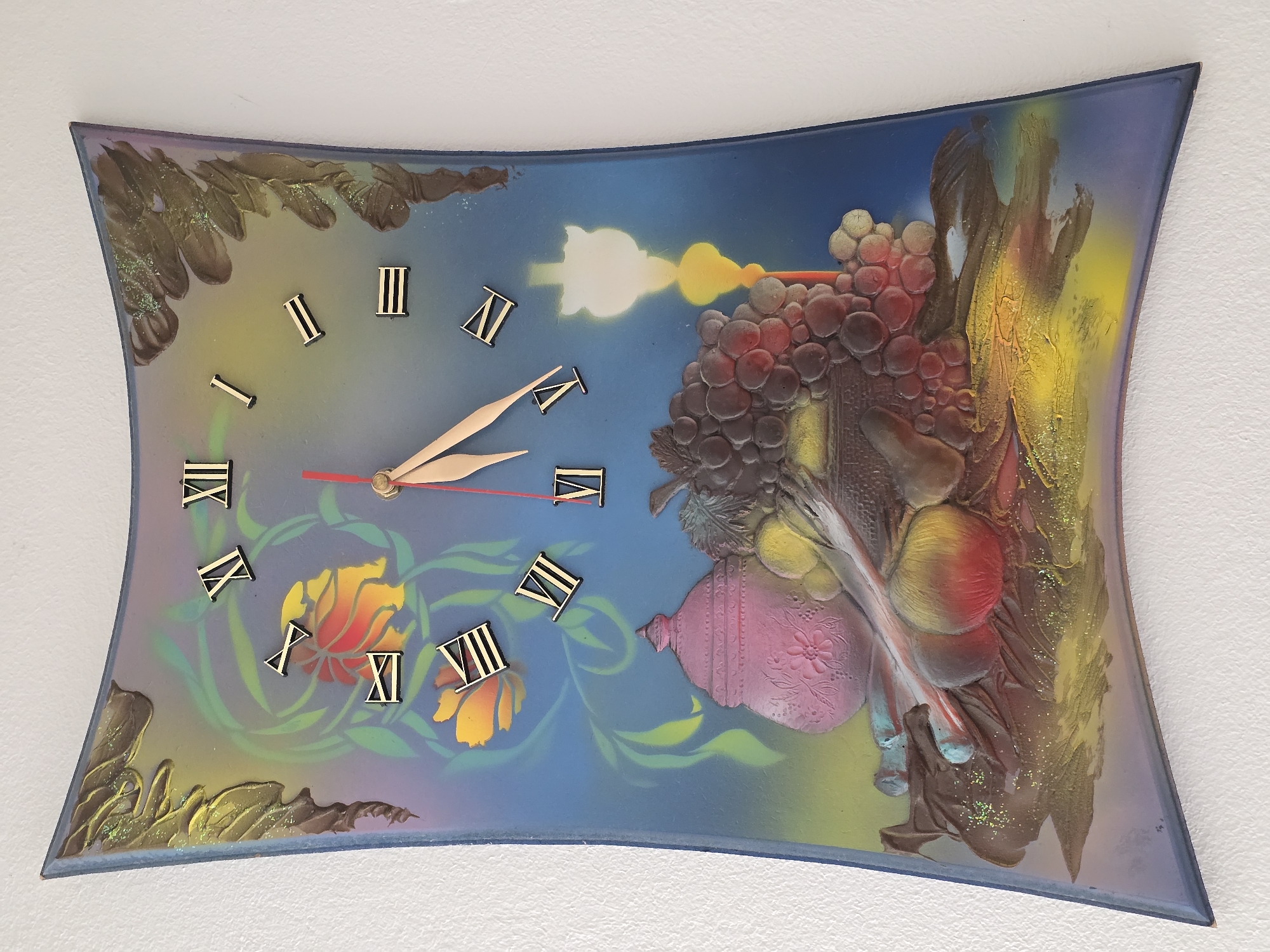 This is an image of a decorative wall clock shaped like a curved rectangle. The clock face is painted with a still life scene featuring a pink pot, grapes, apples, and a candle, all rendered in a somewhat stylized manner. The background transitions from blue to yellow and green, with painted floral elements. Roman numerals mark the hours. The clock hands, one red and one gold, indicate the time. The clock is hanging on a white textured wall. The location of this clock in Herrenberg, Germany, is not determinable from the image.