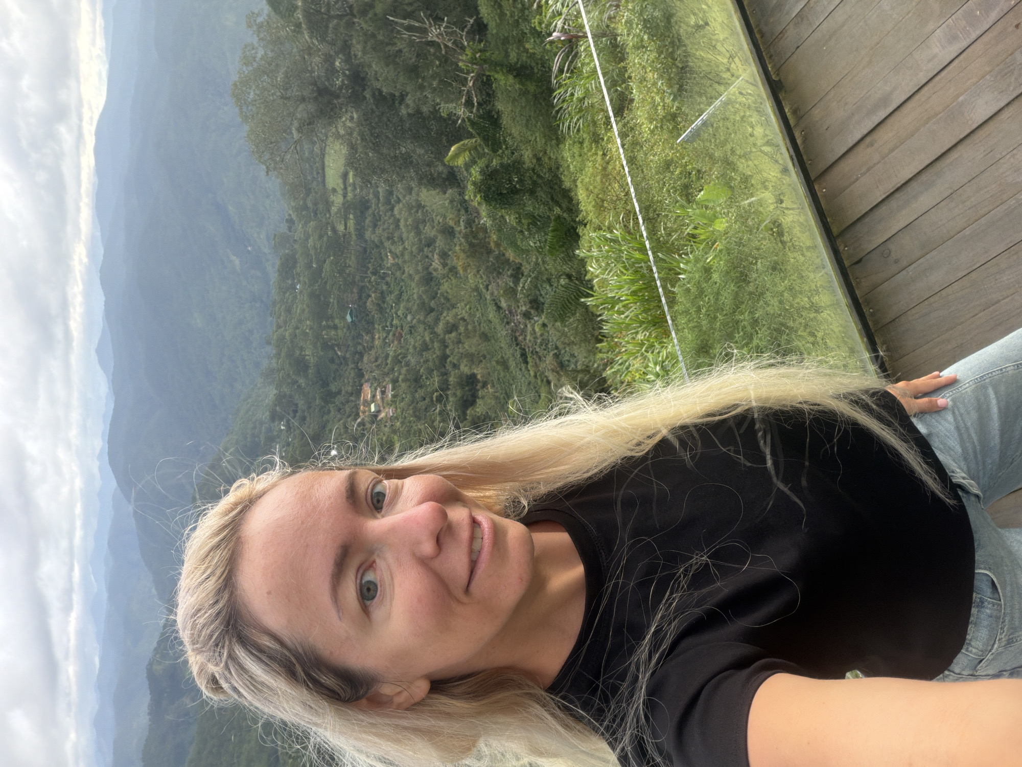 In a selfie taken from the chest up, a blonde woman with fair skin and light blue eyes smiles slightly at the camera. She is wearing a black t-shirt and blue jeans. Her hair is long and blonde, pulled back from her face. She is standing on a wooden deck in front of a glass railing. Behind her, a lush, green valley stretches out, covered in dense trees and foliage. In the distance, rolling hills fade into a hazy blue sky. The setting appears to be mountainous, with layers of hills receding into the background. This scene is located in the city of Phát Chi, Vietnam, which is known for its scenic mountainous landscapes.