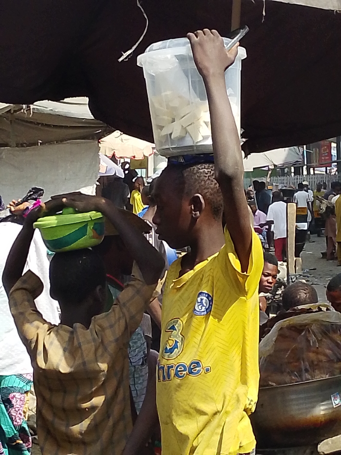 The image depicts a crowded outdoor market scene in Sabon Gari East, Nigeria. A dark fabric canopy partially covers the upper portion of the frame. In the immediate foreground, two young males are prominent. The individual on the right, wearing a yellow short-sleeved shirt with a blue Chelsea FC crest and "3 three." logo, carries a clear rectangular plastic container filled with numerous white, uniformly shaped trapezoidal or rectangular items on his head. His right arm is extended upwards, with his hand resting on the container's lid, holding a small dark object. To his left, a second younger male, wearing a light brown plaid long-sleeved shirt, carries a green and blue lidded plastic bowl on his head, stabilizing it with both hands.
The background is populated with multiple blurred and partially visible individuals, suggesting a busy pedestrian thoroughfare or market area. Structures consistent with market stalls, including white tarpaulins, fabric awnings, and wooden supports, are discernible. In the lower right corner, a large metallic bowl contains dark, possibly fried or cooked food items, partially covered by a clear plastic bag. The scene is brightly lit, indicating daytime conditions.