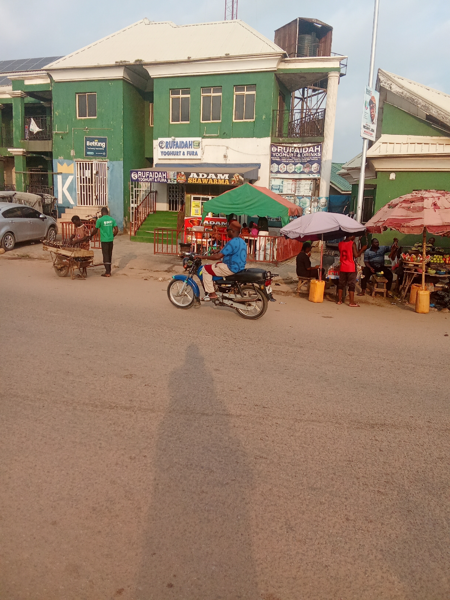 This is a street scene in Kubwa, Nigeria. A blue motorcycle with a rider is traveling on the right side of the road. In the background, a green building with white trim has several windows and a balcony. Signs for "RUFAIDAH YOGHURT & FURA" and "ADAM SHAWARMA" are visible on the building. To the left of the building, two men are standing near a cart with food items. Several market stalls with umbrellas are set up along the sidewalk. A silver car is parked to the far left. The ground is paved with asphalt.