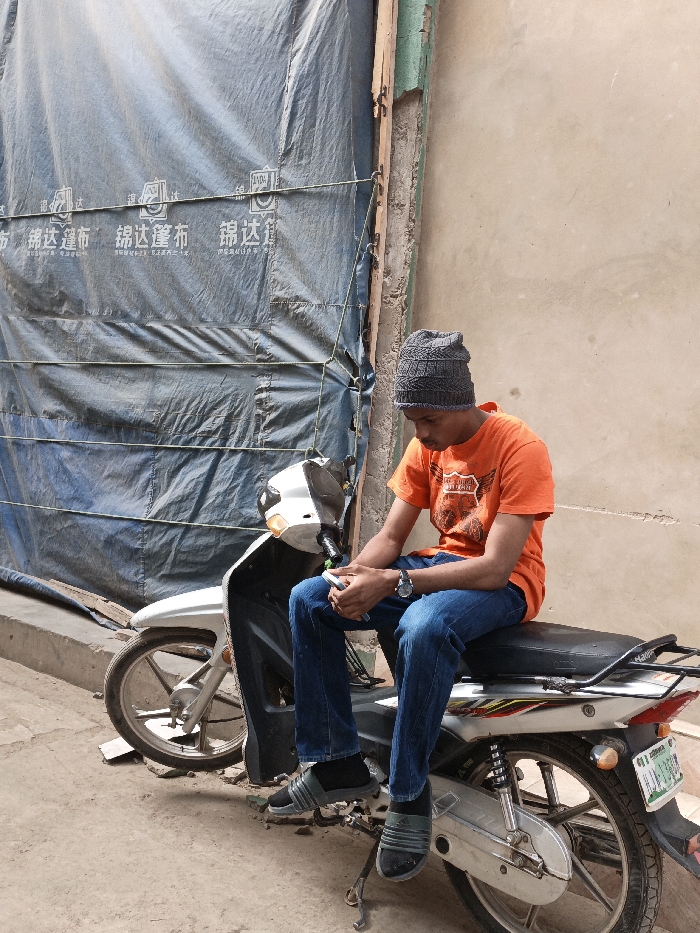 The image displays a young adult male seated on a stationary silver and white scooter-style motorcycle on a concrete surface in Fagge C, Nigeria. The male, positioned facing slightly left, wears a grey knitted beanie, an orange short-sleeved t-shirt with a graphic print, blue jeans, black socks, and black-and-grey Adidas slide sandals. A silver-toned wristwatch is visible on his left wrist. He holds a light-colored mobile phone in both hands, looking downwards at the device. The motorcycle's rear wheel and kickstand are visible, along with a partially obscured green and white Nigerian license plate. The background on the left is dominated by a large blue tarp or canvas, secured horizontally with light-colored ropes, bearing white Chinese characters, including "锦达篷布" (Jin Da Tarp). To the right, a light brown/beige plastered wall features weathered green paint residue near a vertical wooden or metal support structure. The scene is illuminated by natural daylight.