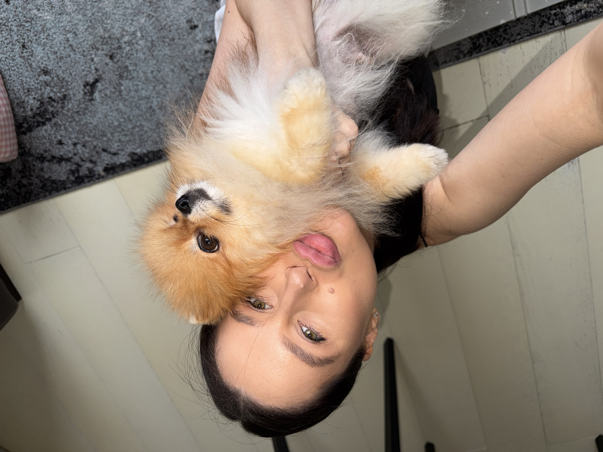 A woman holds a Pomeranian dog close to her face, both facing the camera. The woman has dark hair pulled back, light skin, and is wearing minimal makeup. Her eyes are green. The dog is primarily tan and white, with fluffy fur. The background includes a light-colored wooden floor and a gray carpet. The photo appears to be taken indoors in a domestic setting. There is no evidence in the image indicating the location as Khimki, Russia.