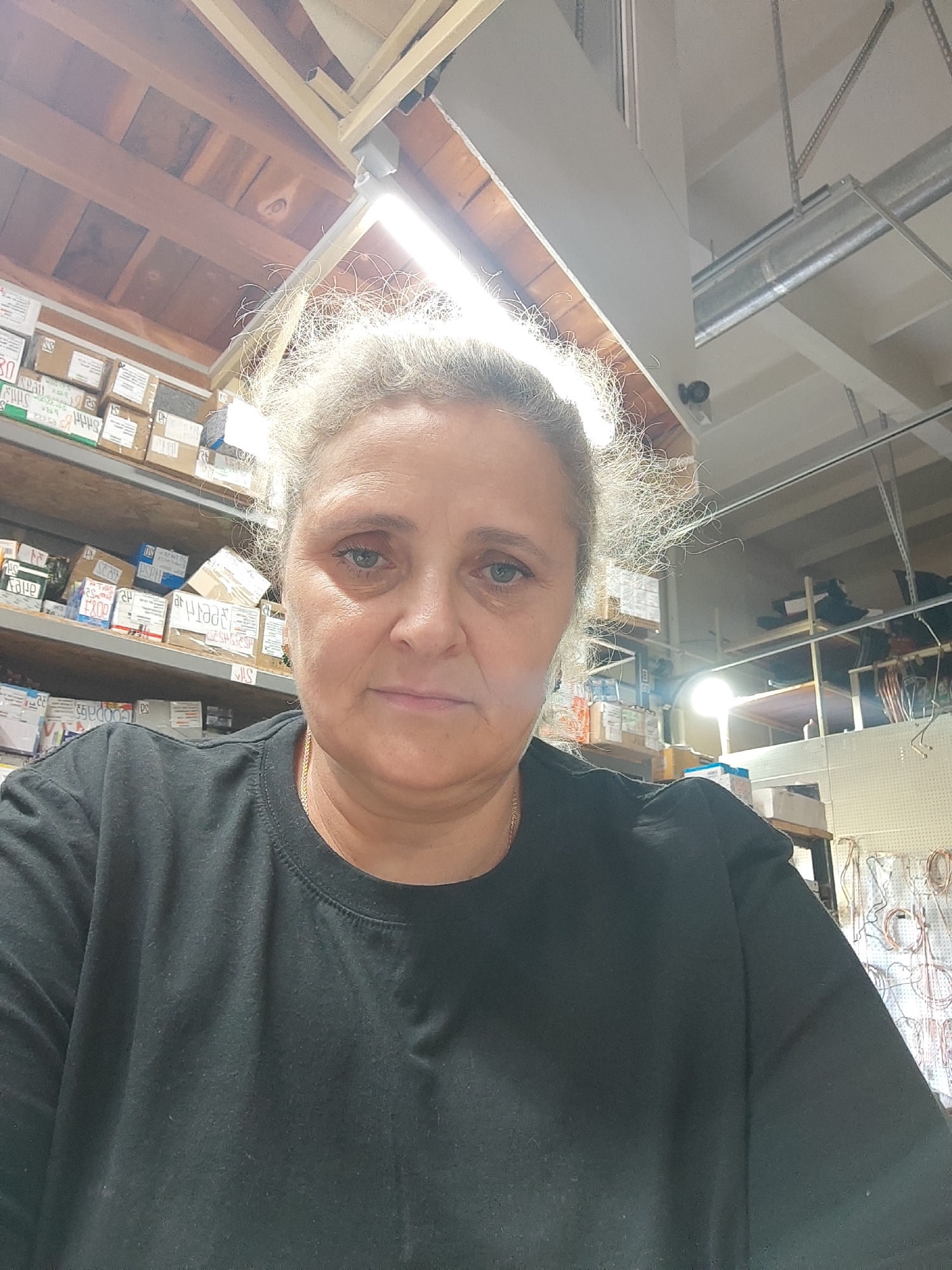 A woman with light-colored, curly hair is shown from the chest up, looking directly at the camera. She is wearing a black t-shirt and a thin gold necklace. Her eyes are open and her expression is neutral. The background appears to be a warehouse or storage area with shelves stocked with boxes. The lighting is bright and appears to be coming from above, possibly fluorescent lights. The overall scene is indoors.