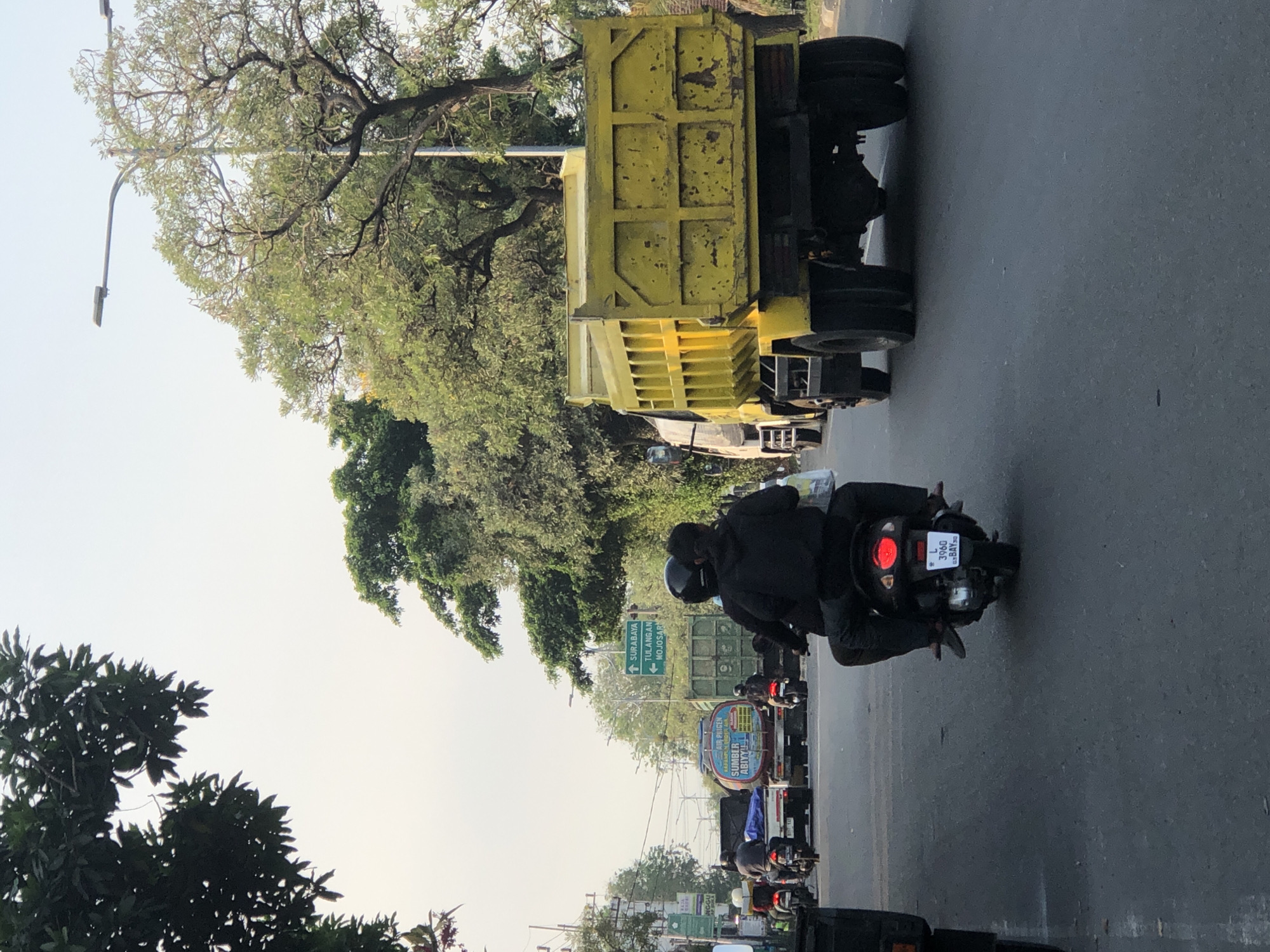 The image captures a street scene in Kludan, Indonesia, with a focus on vehicles and the urban environment. In the foreground, a scooter is seen from behind, carrying two individuals wearing dark clothing and helmets. The scooter's license plate reads "L 3960 G2 BAY50". Further down the road, a bright yellow dump truck is prominently displayed, also viewed from the rear. Other vehicles, including motorcycles and trucks, are visible in the middle ground, some with visible signage such as "AIR PRIGEN SUMBER ABYYYL" and a directional sign pointing towards "SURABAYA", "TULANGAN", and "MOJOSARI". The street is paved, and trees line both sides of the road, with some foliage extending over the sky. The sky is overcast with a pale, hazy hue, suggesting daylight hours. The overall impression is a typical daytime traffic scene in an Indonesian city.