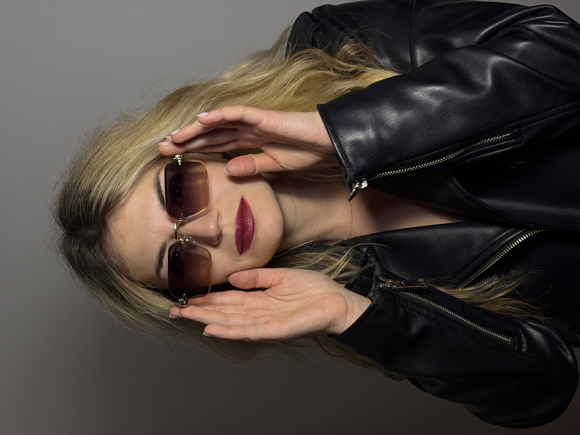 A woman with long blonde hair, wearing a black leather jacket and sunglasses with brown lenses, poses against a plain gray backdrop. She is holding the sunglasses with both hands, positioning them on her face. Her lips are painted with a dark red lipstick. The woman's location is not discernible from the image. Budva, Montenegro, cannot be determined.