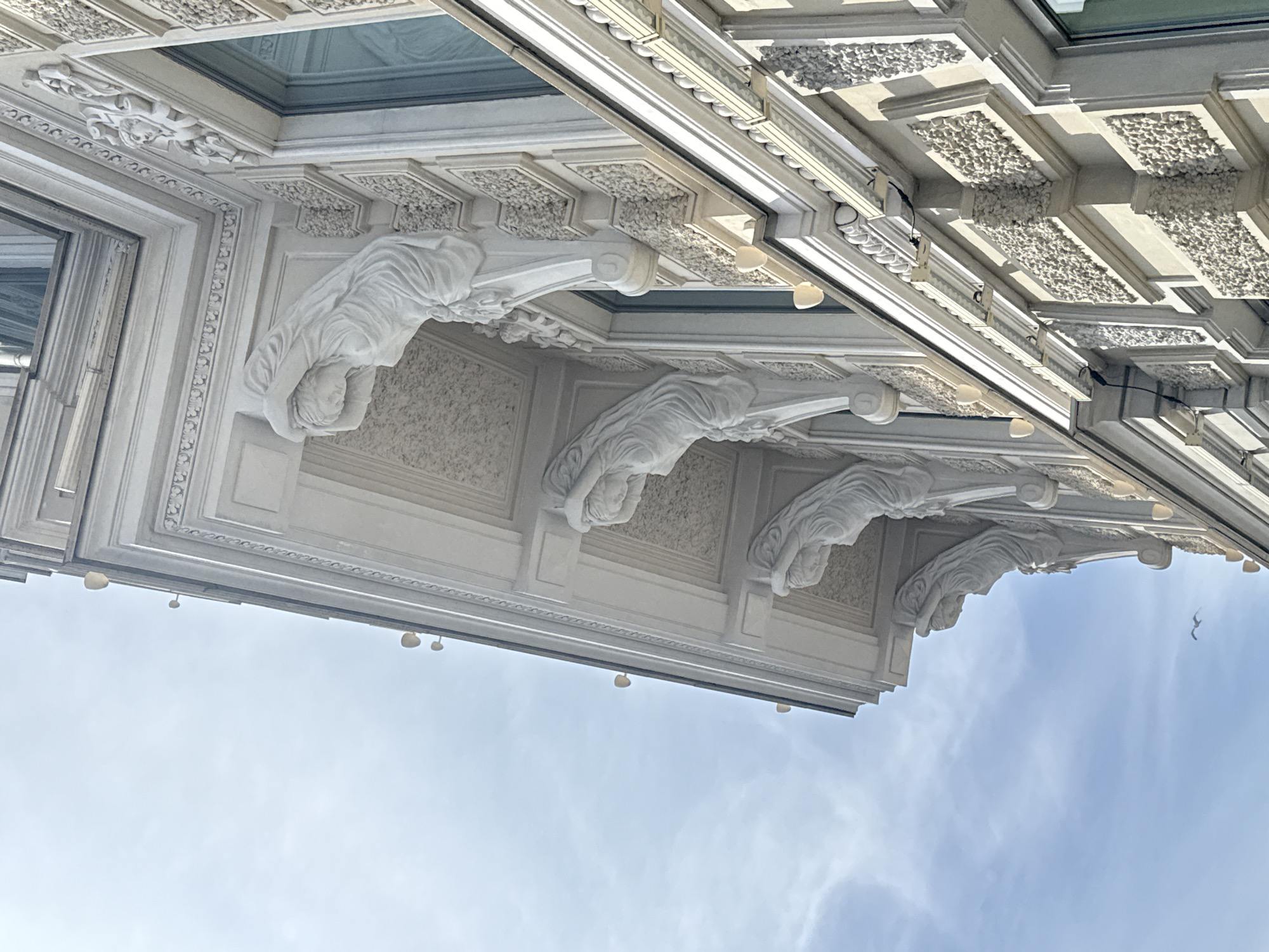The image shows an upward view of an ornate building facade against a pale blue sky with light cloud cover. Architectural details include relief sculptures of draped figures resembling caryatids or atlantes, decorative panels with textured infill, and intricate moldings. The building is white or light-colored, suggesting it may be made of stone or stucco. There are small light fixtures attached to the underside of the building. The architectural style is classical or neoclassical. The photo was taken in Saint Petersburg, Russia.