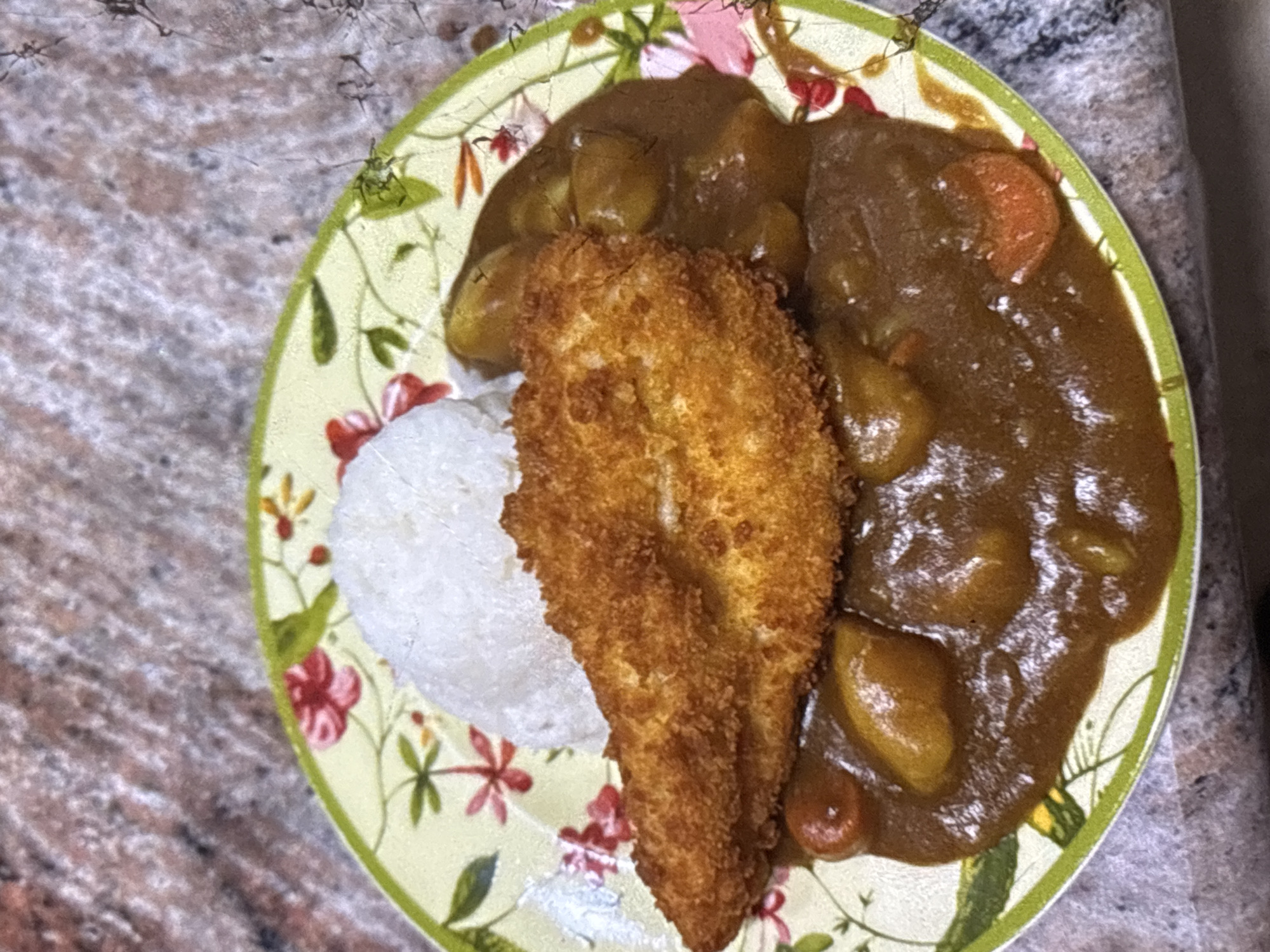 A close-up, overhead shot features a plate of Japanese curry with rice and a breaded chicken cutlet. The plate is decorated with a floral pattern. The curry sauce, a rich brown color, covers the bottom half of the plate and contains visible pieces of potato and carrot. A mound of white rice sits to the left of a golden-brown, crispy breaded chicken cutlet, which is positioned on top of the curry. The background is out of focus, showing a speckled countertop. There is no indication of the city of Orihuela, Spain.
