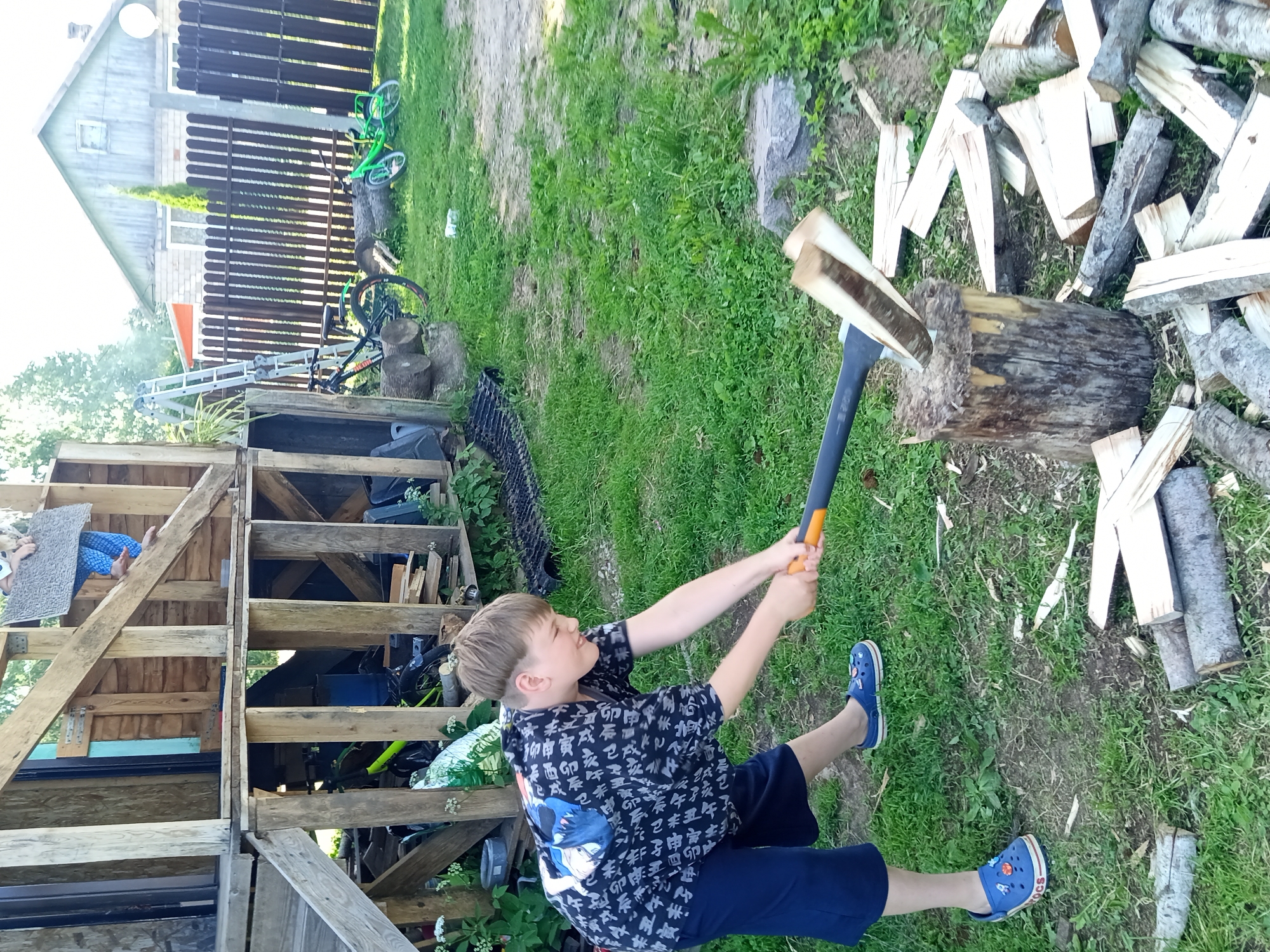 In an outdoor scene in Ostrovtsy, Russia, a young boy is actively engaged in chopping wood. He is wearing a black shirt with a repeating pattern of Asian characters and a blue anime-style graphic on the back, along with navy blue shorts and blue Crocs. The boy is holding a splitting axe with an orange handle and a dark grey head, which is poised to strike a log that is already partially split and resting on a larger chopping block. The ground around the chopping block is covered with a scattering of freshly chopped wood pieces of various sizes, some appearing to be kindling.
The setting appears to be a backyard or a rural area, with a wooden structure, possibly a shed or a multi-level wooden construction, visible to the left of the boy. Behind the wooden structure, a dark brown wooden fence runs horizontally. Two bicycles, one green and another black, are leaning against the fence. The ground is a mix of dirt and green grass, with some patches of dry soil. In the background, a light-colored house with a dark roof is visible. The lighting suggests it is daytime, likely a sunny day given the brightness and the shadows. The overall mood seems to be one of focused activity, possibly a chore or a recreational task. No specific text is legible in the image.