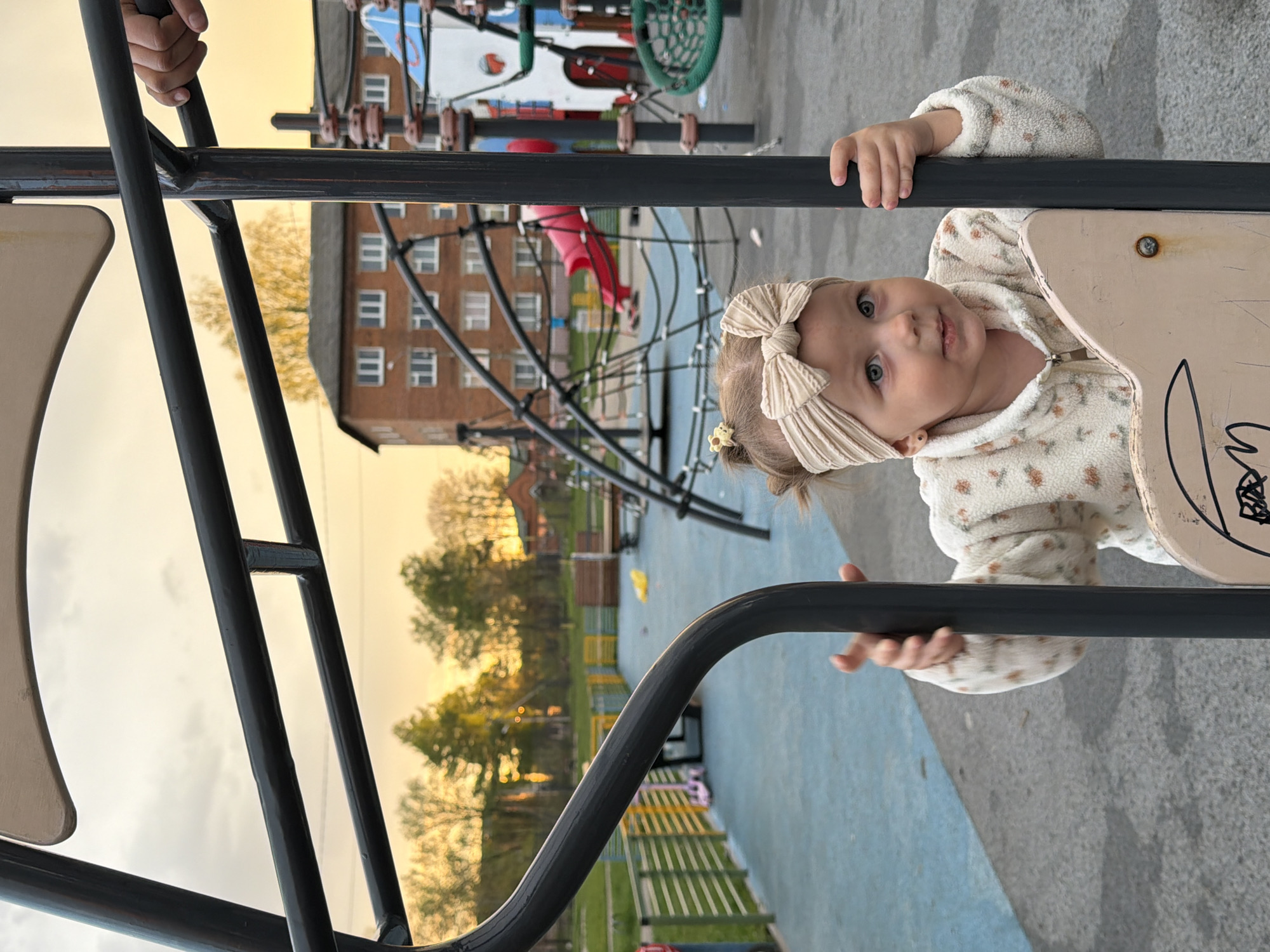The image shows a child with a light-colored bow in their hair, wearing a floral print jacket, holding onto a metal structure, possibly part of a playground in Reshetnikovo, Russia. In the background, there's a multi-story brick building, trees, and other playground equipment. The light suggests it's either sunrise or sunset. The ground is a mix of blue and grey, potentially rubberized flooring. A hand of another individual is seen at the top of the metal bars on the left.