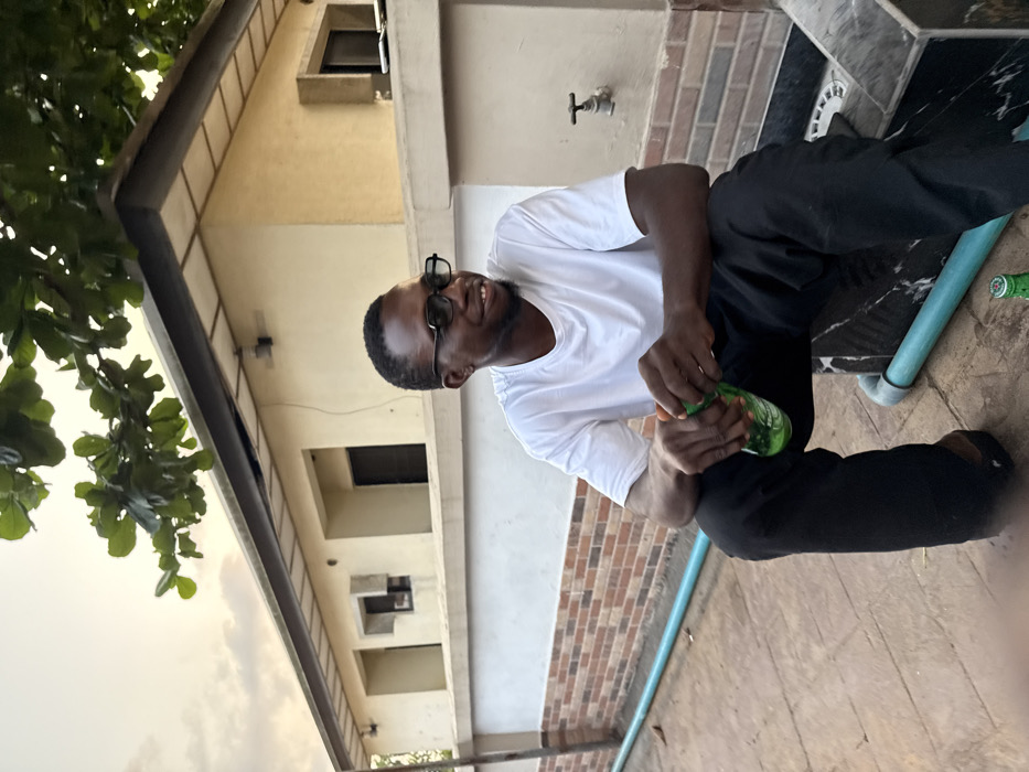 The image displays an outdoor scene in Badore, Nigeria, depicting a single adult male during late afternoon or early evening hours. The individual, dark-skinned with short dark hair, wears a white short-sleeved t-shirt, black trousers, and black-framed glasses. He is seated, oriented slightly to the right of the frame, with a slight smile, and holds a green plastic beverage bottle with both hands, appearing to interact with its cap. Another identical green bottle rests on the reddish-brown tiled ground near his feet, adjacent to a light blue pipe that runs horizontally across the foreground. The male is positioned next to a dark, marble-patterned structure. Behind him, a white wall with a lower section of brown brick patterning features a brass water spigot. In the background, a light-colored building with a dark roof overhang is visible, along with several recessed rectangular window openings. Green tree foliage occupies the upper portion of the frame against a diffused, light-colored sky.