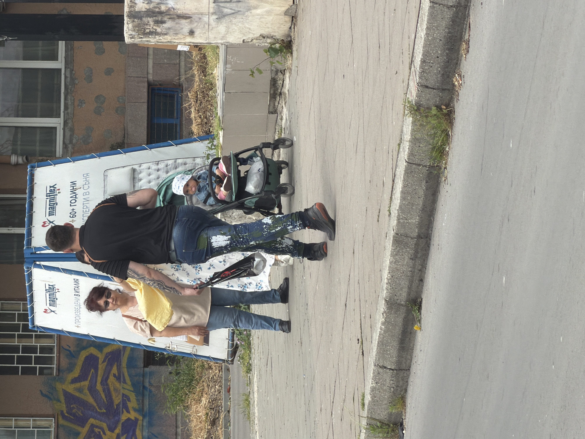 A daytime street scene in Varna, Bulgaria, depicts a man, a woman, and a baby in a stroller on a sidewalk next to a street. The man is wearing a black shirt and jeans with paint splatters. The woman is wearing sunglasses, a tan shirt, and jeans. The baby is in a stroller. The sidewalk is made of concrete. To the left of the people is a sign with the word "magniflex" on it and text in Bulgarian. In the background, there is a building with graffiti on it.