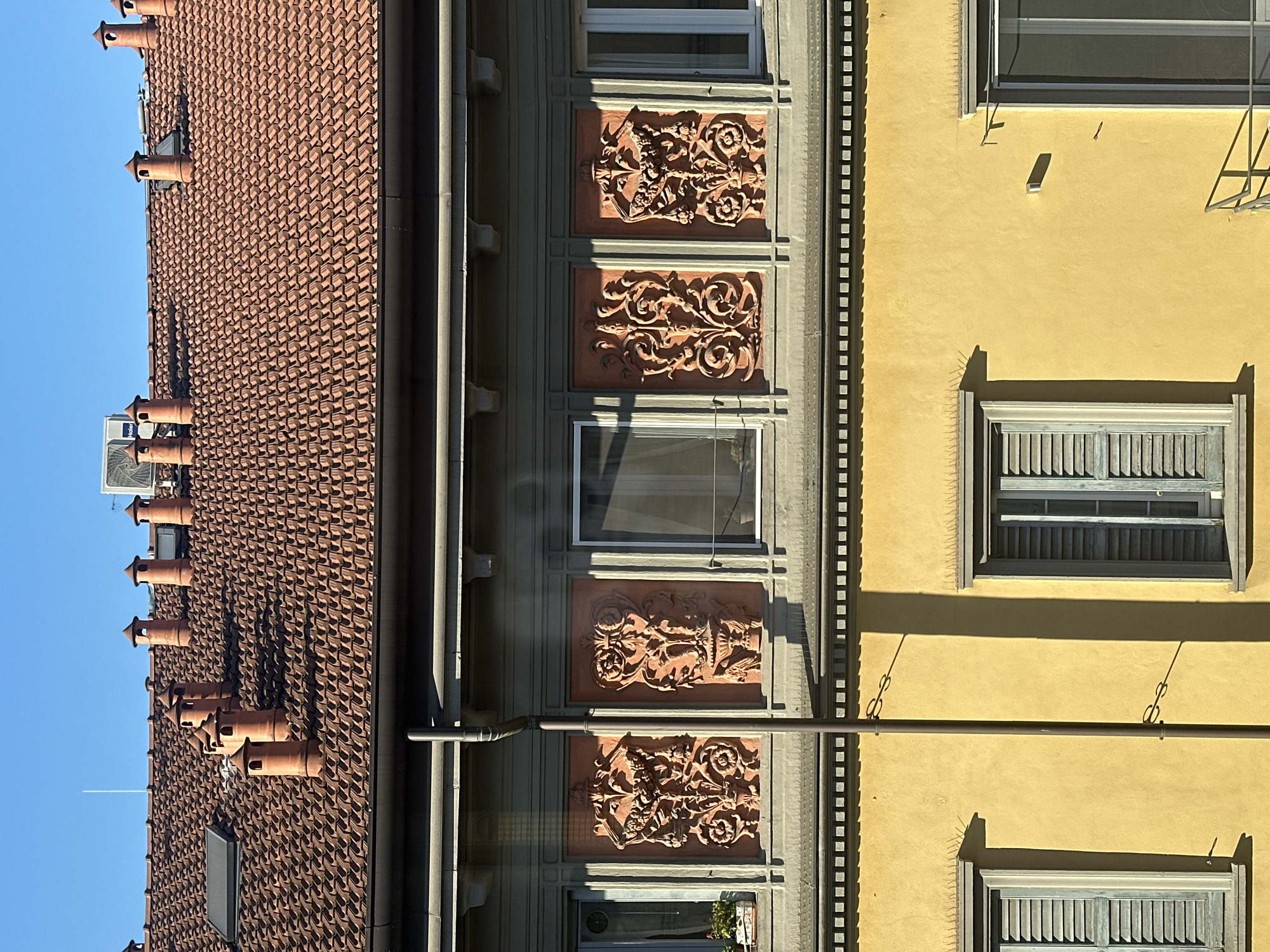 The image shows the exteriors of three buildings in Milan, Italy, under a clear blue sky. The building on the left has a red-tiled roof with multiple orange chimneys, and a visible air conditioning unit. The middle building is painted in a muted green color, featuring ornate reddish-brown decorative panels between windows. The building on the right is yellow and includes windows with blue shutters. A horizontal pipe runs across the facade of the two rightmost buildings.