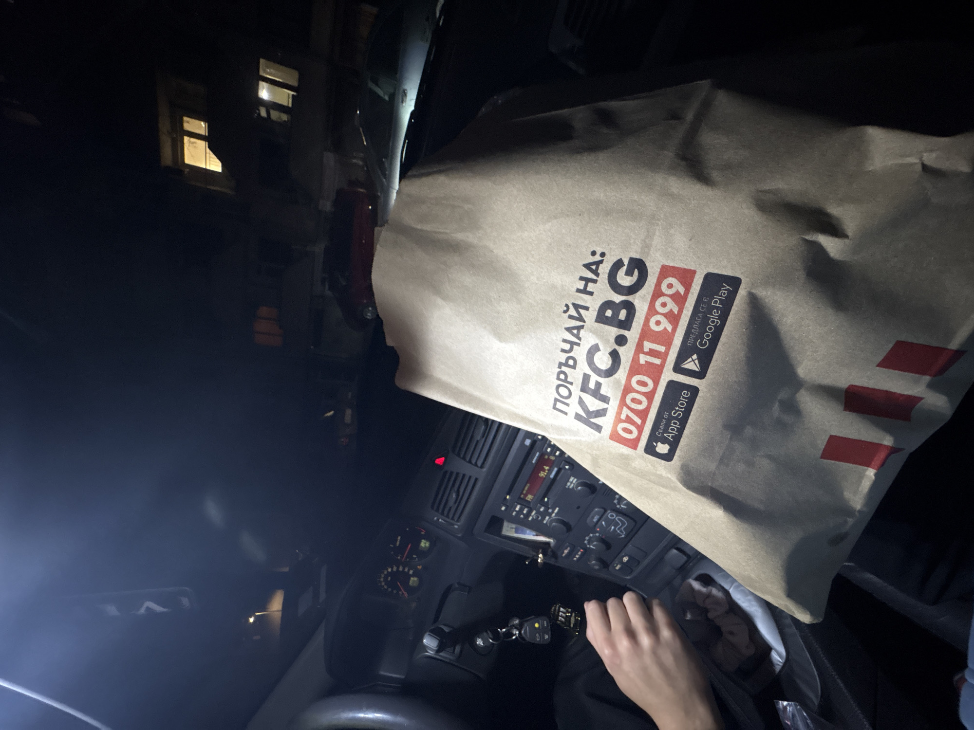 The image captures a nighttime scene inside a car in Plovdiv, Bulgaria, showing a person holding a large KFC paper bag. The bag, centrally positioned and tilted, features the KFC logo and prominent text in Bulgarian: "ПОРЪЧАЙ НА: KFC.BG" (Order at: KFC.BG), a phone number "0700 11 999," and prompts to download the app from "App Store" and "ПРЕДЛАГА СЕ В Google Play" (Available on Google Play).
In the foreground, part of a person's left hand is visible, positioned near the car's gear shift and keys. The car's dashboard is partially visible, featuring a radio displaying "91.4" (likely a radio frequency), air vents, and various buttons and controls. Gauges are also discernible to the left of the dashboard. Car keys with a keyring are hanging from the ignition. A pink scrunchie is placed in a cup holder or storage compartment in the center console.
Outside the car, through the windows, illuminated windows of a building can be seen, indicating the late hour. The overall lighting is dim, with artificial light from inside the car illuminating the immediate foreground and the glow from distant building lights in the background. The image suggests a moment after picking up a food order, possibly for a meal.