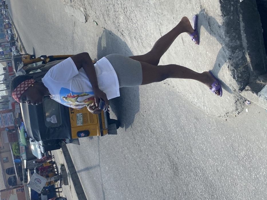 The image captures a female subject standing on a concrete curb next to an asphalt road in Badore, Nigeria, under clear daylight conditions. The subject is a dark-skinned individual wearing a red and white patterned headband, a white short-sleeved t-shirt with a colorful graphic print on the front, and light grey form-fitting shorts. Her feet are clad in purple open-toed slide sandals. She holds a dark, rectangular object, possibly a mobile phone, in her left hand, positioned in front of her. Her head is turned slightly to her right, and her gaze is directed off-frame. Directly behind her, on the asphalt, is a yellow Keke Napep (auto-rickshaw) displaying "TVC" and the license plate "LAGOS GGE-871LC" on its rear. Further down the road, other vehicles and pedestrians are visible. Buildings, appearing to be commercial or residential structures, line the background. The concrete curb shows signs of wear and minor debris. Defined shadows are cast on the ground, consistent with direct sunlight.