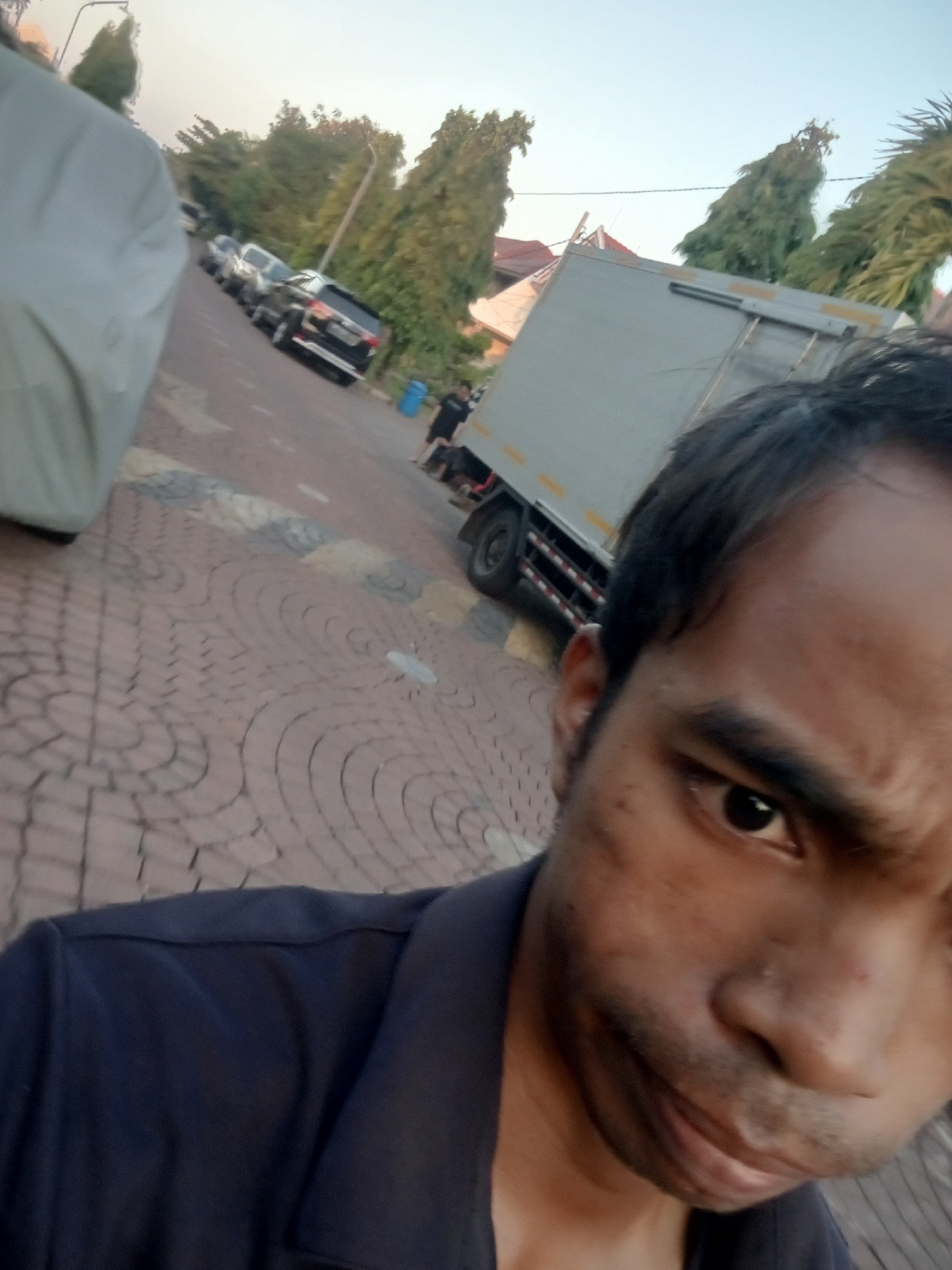 In an outdoor scene, a person with dark hair and an expressive face is captured in a close-up, tilted selfie. They are wearing a dark blue collared shirt. The background reveals a street with parked cars and a large truck, suggesting an urban environment. The pavement is paved with red bricks arranged in a patterned walkway. Trees and buildings are visible in the distance, along with a clear sky. Based on the distinct brick pattern and the presence of the iconic Surabaya city lamp posts, this scene is located on Jalan Tunjungan, a historic street in the city of Surabaya, Indonesia.