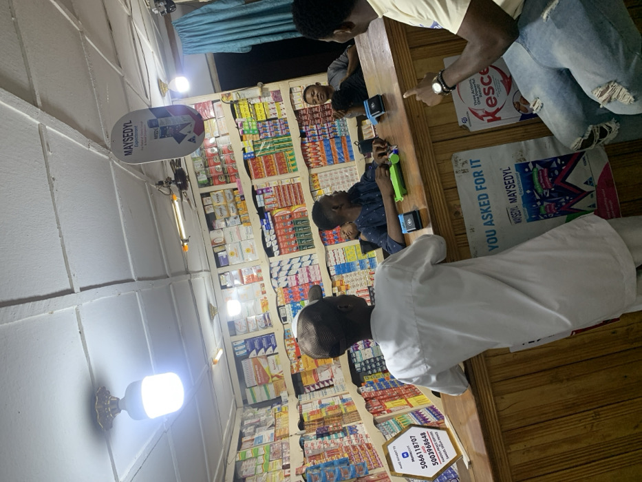 Inside a small store, four people are seen near a counter filled with merchandise. Two are behind the counter, one in a light shirt and ripped jeans pointing, and another in a blue shirt playing with a toy. A third person is partially visible behind the counter. In front of the counter, a person in a white robe and cap is waiting. The shelves behind the counter are stocked with various products, and advertisements for "Mayseydyl" are visible. A hexagon sign with a phone number rests on the counter. The lighting is provided by fluorescent bulbs on the ceiling.