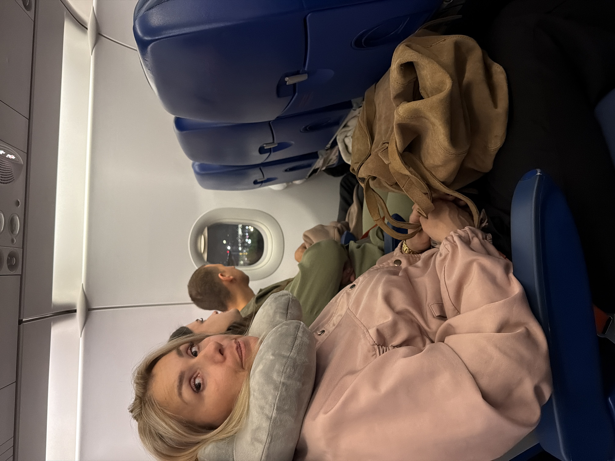 The image shows the interior of an airplane cabin, with a woman in the foreground looking at the camera. She is wearing a pink jacket and has a neck pillow around her neck. Next to her, a brown suede bag rests on her lap. Behind her, a man is looking out the window, which offers a view of city lights at night, suggesting Saint Petersburg, Russia. Several rows of blue airplane seats are visible in the background. The overall scene depicts passengers on a nighttime flight.