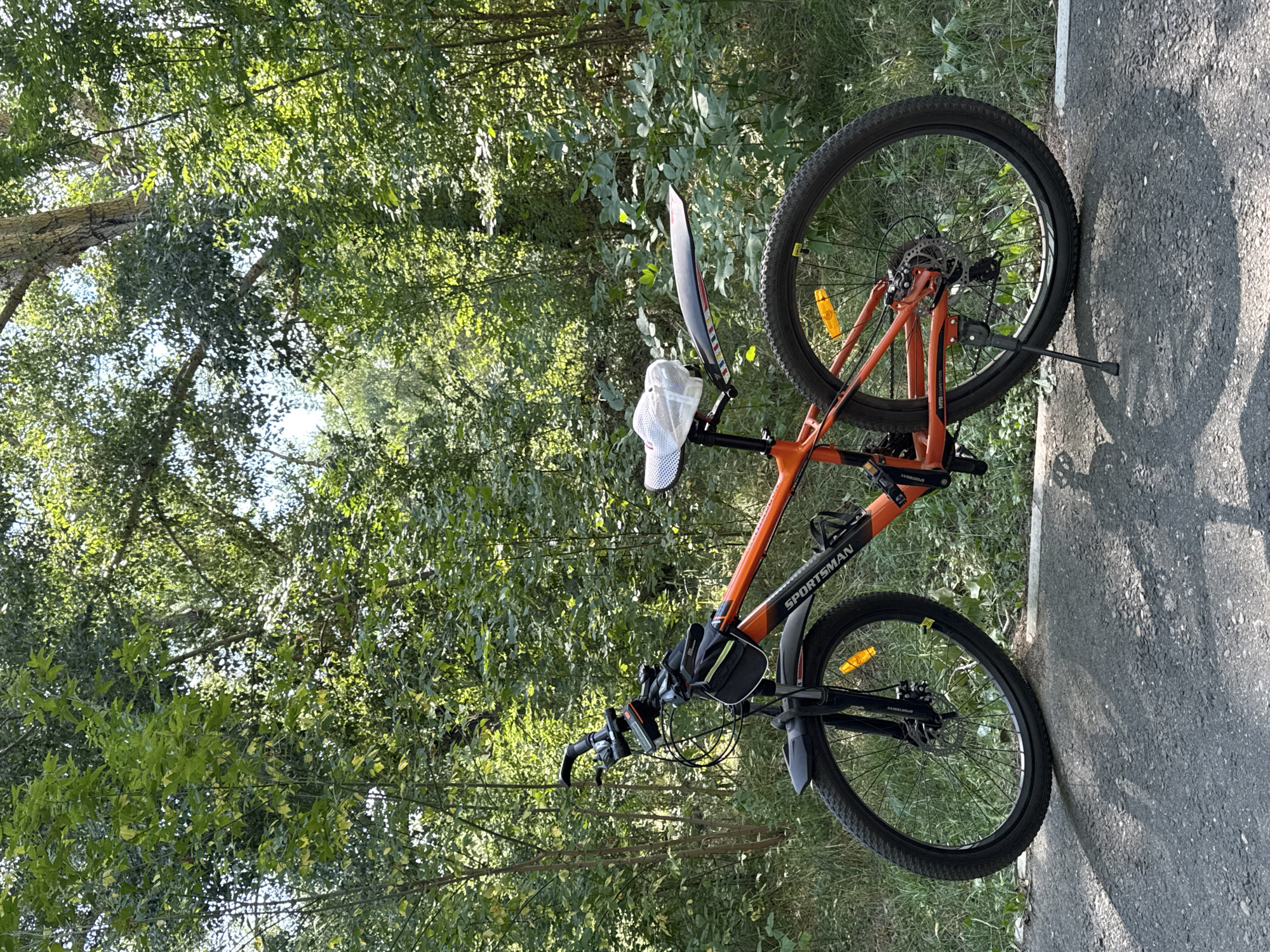 An orange and black mountain bike is parked on a paved surface next to a grassy area, with a dense forest in the background. The bike has a black saddlebag attached to the handlebars and a white mesh cover over the saddle. The front wheel has a black fender and a yellow reflector. The rear wheel has a black fender with a colorful striped decal and a yellow reflector. The bike has a kickstand. The environment appears to be a park or recreational area in the city of Oskemen, Kazakhstan, characterized by lush green trees and foliage, with sunlight filtering through the leaves. There are no people present in the image.