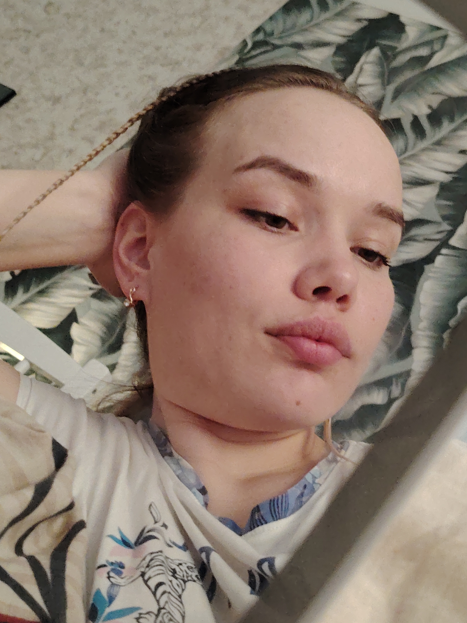 A close-up, angled portrait features a young woman with fair skin and dark brown hair, styled with fine braids on the side of her head. She has defined eyebrows, dark eyes, and full lips, looking downwards and slightly to her right with a calm, neutral expression. She is wearing small gold-toned hoop earrings with a white bead in her visible right ear.
Her attire includes a light-colored t-shirt or top featuring a detailed print of a black and white tiger along with blue and green tropical leaf motifs. She appears to be reclining or resting, with her right hand gently placed behind her head. The background consists of a patterned pillow or blanket with large green tropical leaves on a lighter fabric. A blurred, light-colored object, possibly a piece of furniture, is visible in the bottom right foreground. The lighting is soft, suggesting an indoor setting. There is no visible text in the image. The provided context indicates the location is Марковский, Russia.