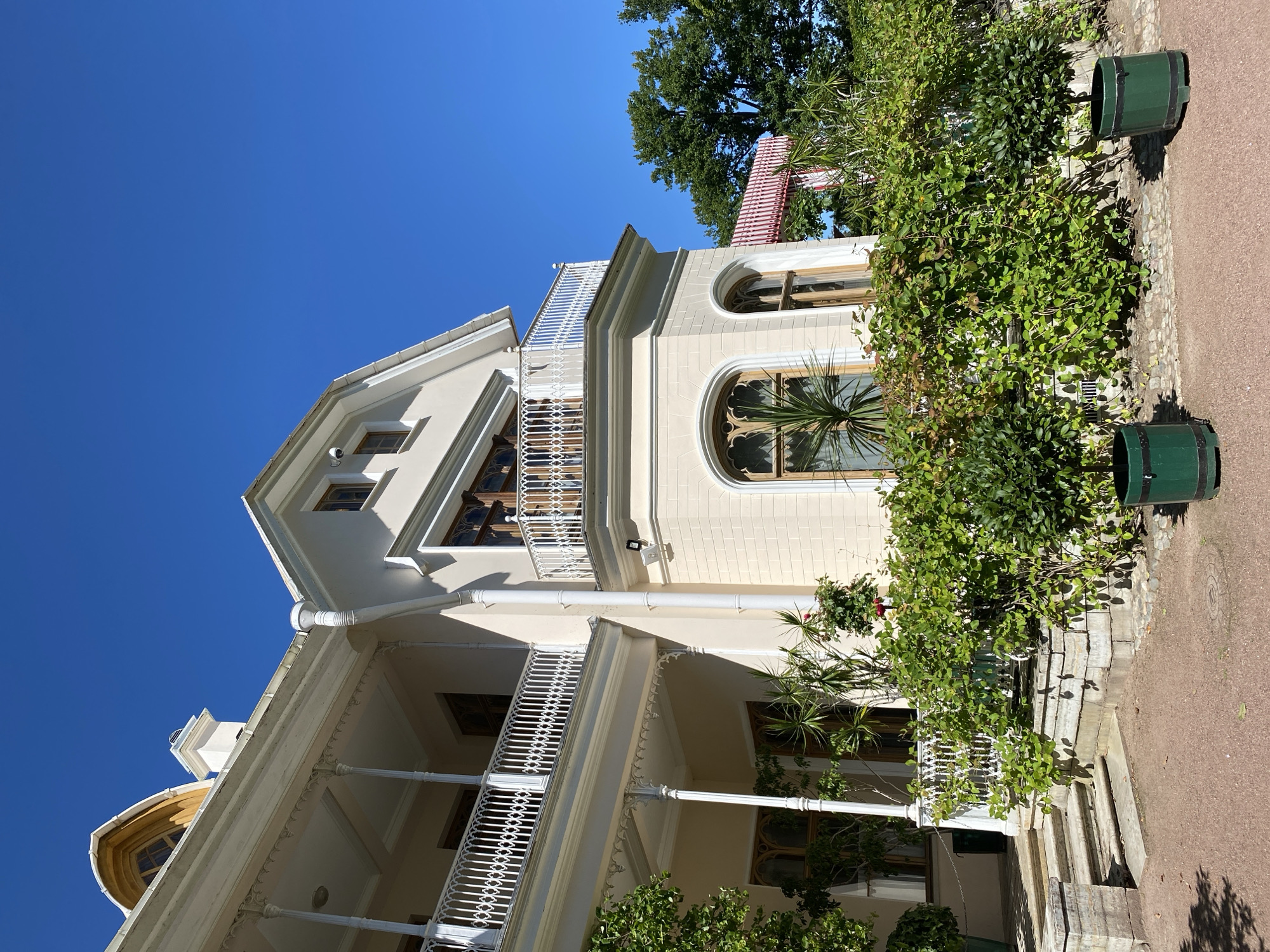 This is a low-angle, full shot of a cream-colored building with ornate white railings on its balconies. The building has several arched windows with dark wooden frames. Green foliage, including bushes and trees, surrounds the building, with two dark green cylindrical planters in the foreground. A red and white awning is visible behind the foliage. The sky is a clear, bright blue. The building is part of the Alexandria Park in Peterhof, Russia, specifically it is the Cottage Palace.
