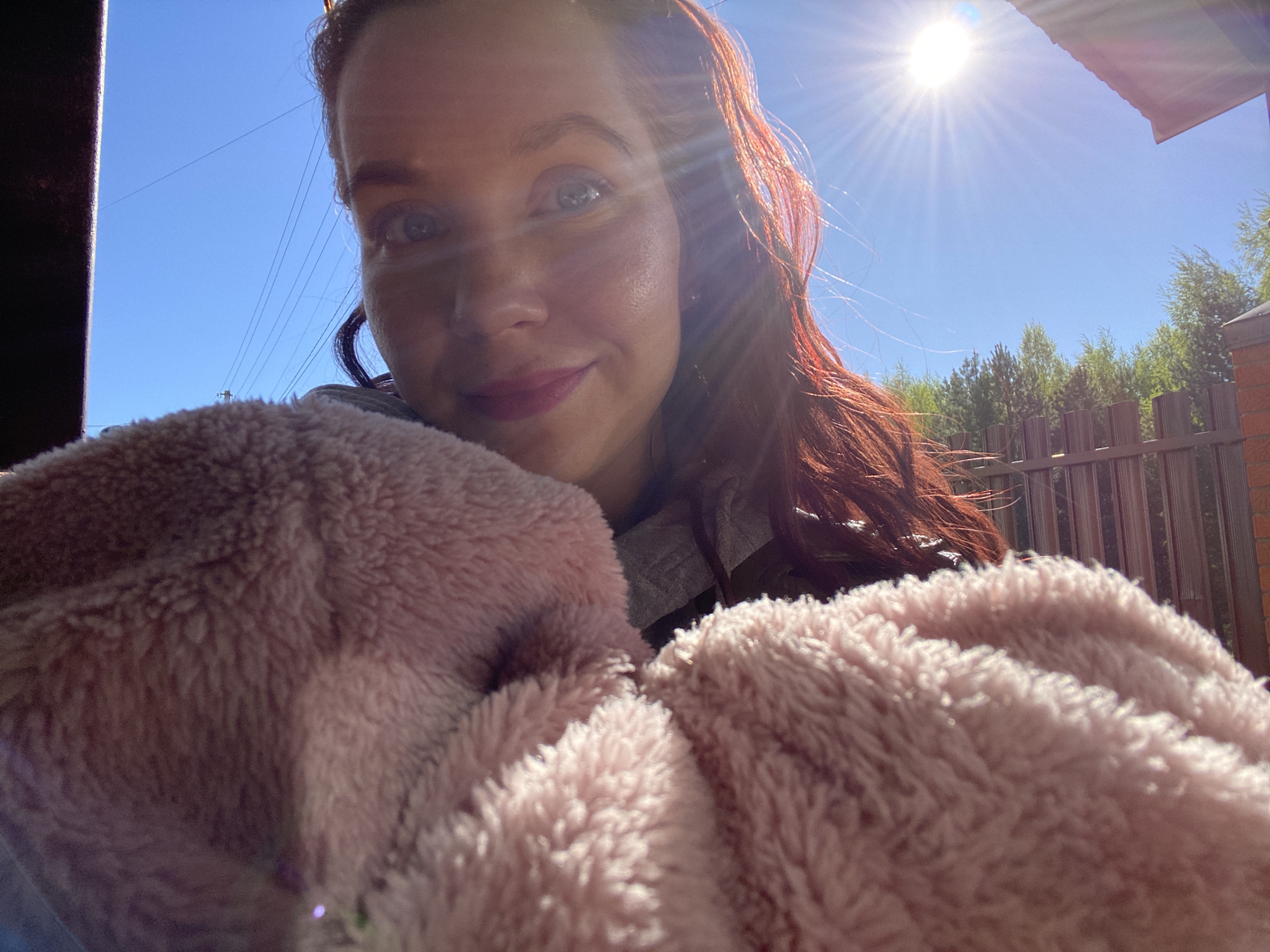 A selfie shows a fair-skinned woman with reddish-brown hair, a light smile, and visible pores, illuminated by sunlight. She is holding a pink, plush garment or blanket close to the camera, obscuring the lower half of the frame. The background features a clear blue sky with the sun prominently positioned, and hints of a residential area with a brown fence, greenery, and parts of buildings or structures. Electric lines are also visible in the sky. The location is in Фоки, Russia.
