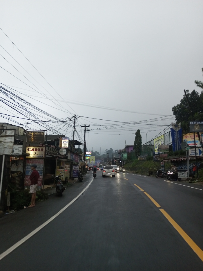 In the city of Dawathang_Dorjibi_ Kashingtsawa, Bhutan, an outdoor street scene is depicted under an overcast, bright gray sky, with atmospheric mist or fog obscuring distant hills. The asphalt road, wet from precipitation, extends towards the background, marked by a solid white line on the left and a broken yellow centerline. On the left side of the road, a male individual wearing a red long-sleeved shirt, light-colored shorts, and a cap stands barefoot on a sidewalk adjacent to a row of commercial buildings. These structures display various signs, including "Canon Digital Copier," "Beauty Make Up," and "ATM." A dense network of utility wires spans overhead, connected to multiple utility poles along the street. Several motorcycles are parked on the sidewalk along the left side. In the mid-ground, vehicular traffic is present on the wet road; a white compact car with illuminated headlights is observed traveling away from the viewer, preceded by a motorcycle. Additional vehicles with headlights on are visible further down the road. The right side of the street features an elevated bank with more commercial signage, including partial views of "RESTORAN" and "VILLA APAYA," and a tall, slender evergreen tree. Several motorcycles are also parked on the shoulder along the right side. The overall lighting suggests either dusk or a heavily overcast day.