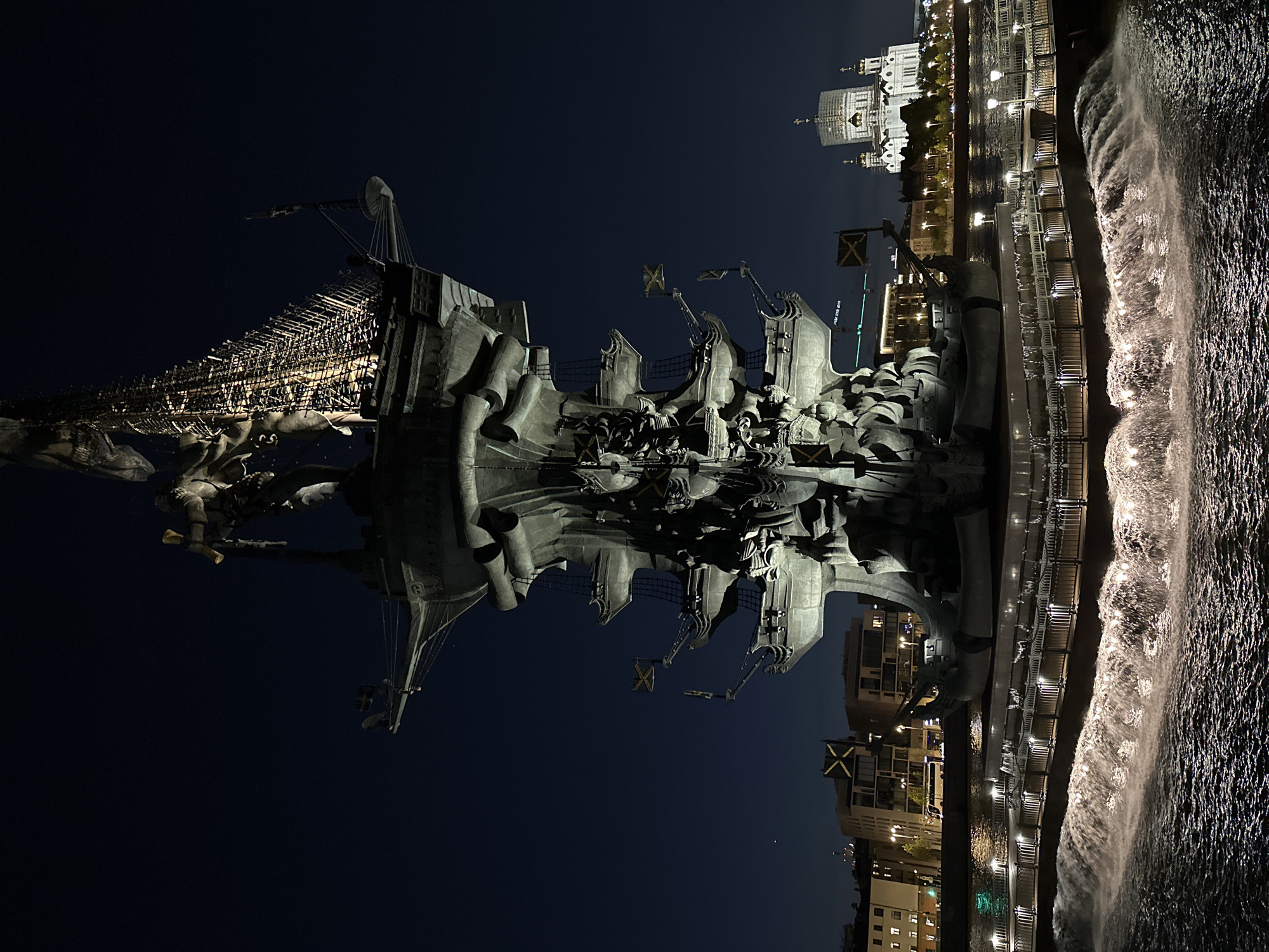 At night, the camera captures a vertical shot of the illuminated monument to Peter the Great in Moscow, Russia. The statue, which is rendered in dark metal, depicts a ship with sailors and is adorned with flags featuring the cross of Saint Andrew. It casts a strong reflection on the water below. Adjacent to the monument is a sidewalk lined with lights, and the cascading water feature is visible on the right side of the frame. In the background, buildings are illuminated. The Cathedral of Christ the Savior is visible on the upper right side of the frame. The overall ambiance of the image is dark and moody.