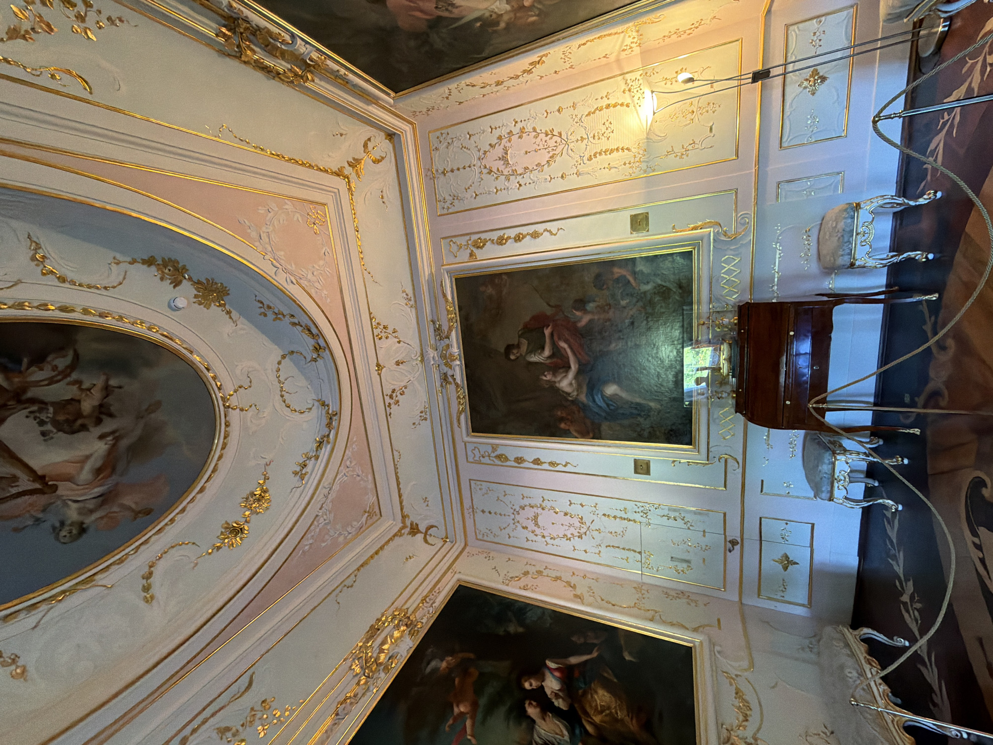 An ornate room in a historic building features a domed ceiling with a fresco, gilded decorative elements, and ornate wall paneling. Two large paintings are prominently displayed on the walls, one on the left depicting figures in classical attire and another on the right with a similar theme. In the center of the room, a dark wood desk with brass accents sits between two small, upholstered stools. A floor lamp with a curved neck stands to the right of the desk. The floor is dark wood with a decorative inlaid pattern. Velvet ropes are placed around the furniture and paintings, suggesting the room is part of a museum or historical site. The lighting is subdued, highlighting the rich details of the interior design. The room appears to be empty of people.