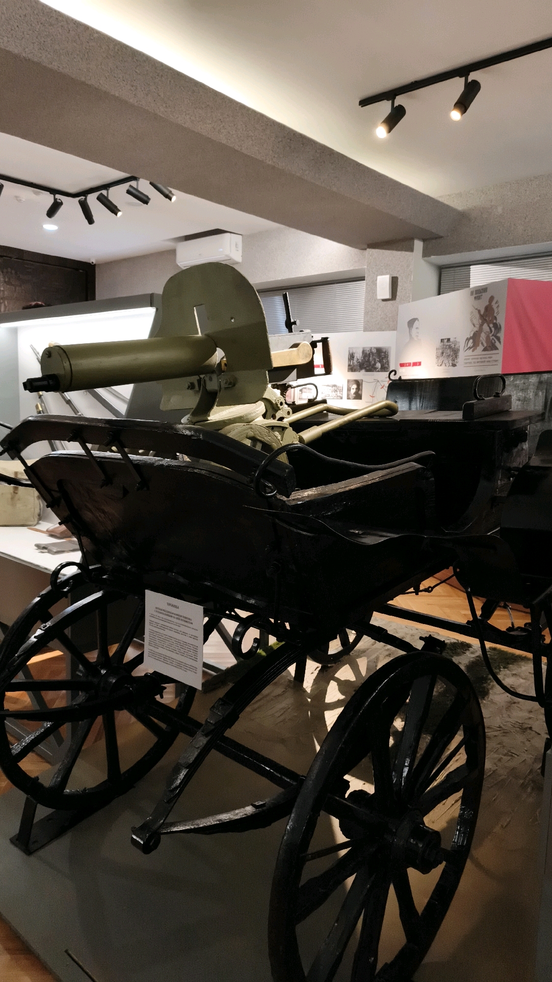 An antique machine gun, painted olive green, is mounted on a horse-drawn carriage within a museum setting. The carriage, made of dark wood with black metal accents and spoked wheels, appears to be a historical artifact. A sign with text is visible on the side of the carriage. The background shows museum walls with framed historical images and displays, and overhead lighting fixtures. The floor is made of polished wood. The machine gun has a long barrel and a complex mount. The overall impression is that of a historical exhibit showcasing early military technology. The location is the Museum of Military Glory in Cheboksary, Russia.
