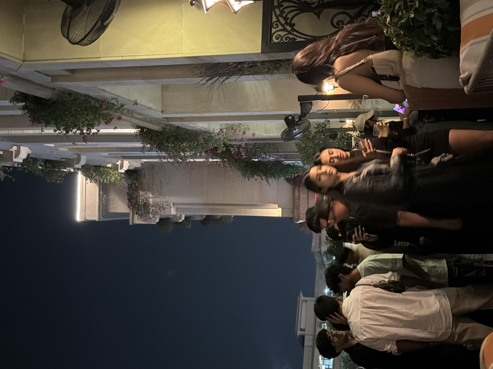 The image depicts a nighttime social gathering at an upscale outdoor restaurant or lounge within a commercial or entertainment district in New Delhi, India.
The background features a multi-story building with a light-colored facade, characterized by repeating columns, ornate balconies, and decorative ledges illuminated by strip lighting. Lush green foliage and pink flowering plants cascade from these balconies, adding natural elements to the urban structure. The sky is dark blue, indicating evening hours. To the far left, another building with a flat roofline is partially visible against the night sky. Several wall-mounted fans, dark in color, are visible on the building's facade, positioned to provide air circulation. On the far right, a section of an intricate dark metal railing or gate with scrollwork patterns is visible, beside which hangs a decorative, star-patterned lantern emitting warm light.
In the midground and foreground, approximately ten individuals are congregated, primarily engaged in conversation. On the left, a cluster of men stands, some holding drinks. One man in a white shirt and light-colored trousers, seen from the back, is central to this group, while another man with glasses smiles towards a third individual. In the center-right, two women stand prominently. One woman with long dark hair, wearing a black outfit, looks towards the left. Beside her, another woman with long dark hair holds a smartphone in her left hand and a drink in her right. Behind them, a person wearing a light-colored baseball cap holds a drink. Further to the right, a woman with long brown hair, seen from behind, wears a light-colored, possibly off-white, dress or top and carries a matching shoulder bag. She is positioned at a dark stand, appearing to interact with a tablet or screen mounted on it. In the bottom right corner, a section of an orange and white striped upholstered chair or bench is visible. The scene is illuminated by warm artificial lighting, creating a lively atmosphere.