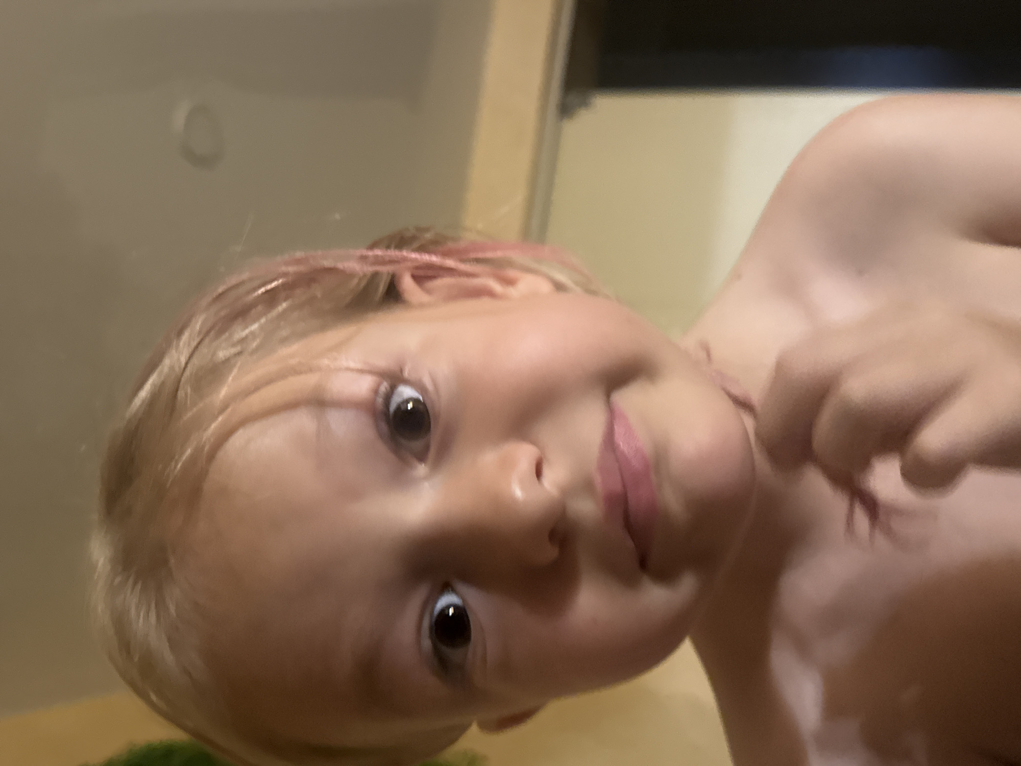This is a close-up, low-angle shot of a young child's face. The child has blonde hair with streaks of pink. They are looking directly at the camera with wide, dark eyes and a slight smile. The child's skin appears fair. Part of their shoulder and chest are visible, and they are touching their neck with their right hand. The background is blurred and appears to be an indoor setting with light-colored walls. The lighting suggests it could be daytime, but no specific time can be determined. There is no visible text in the image.