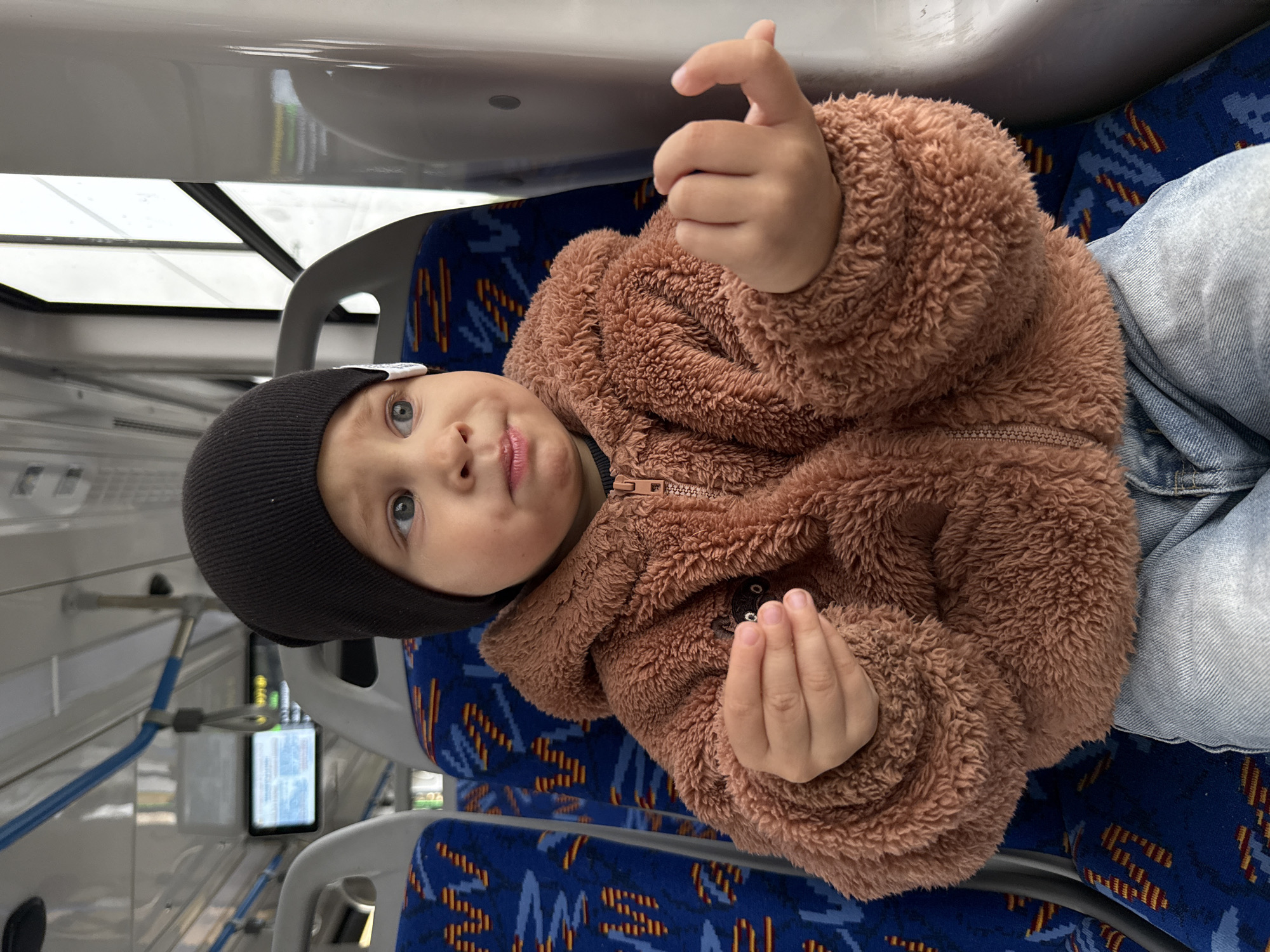 A child is sitting in a bus seat, looking to the right. The child is wearing a black beanie, a brown fuzzy zip-up jacket, and blue jeans. The child is holding something small in their left hand. The bus interior is visible, with blue patterned seats and a window on the right. The location is identified as Rusanovo, Russia.
