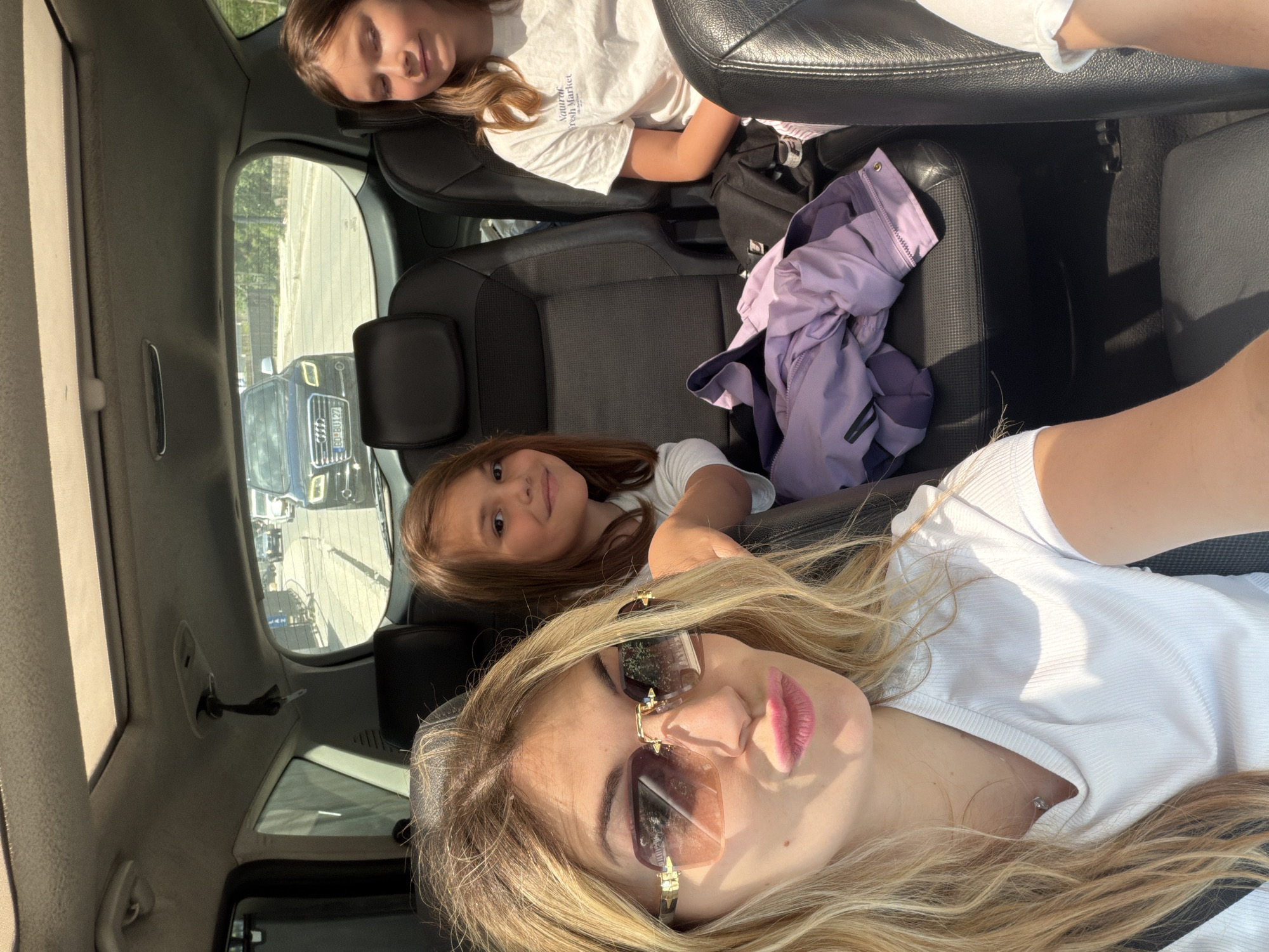The interior of a vehicle is shown with three individuals visible. In the foreground, a blonde woman with sunglasses is taking a selfie. In the middle row of seats, a younger girl with brown hair is looking at the camera. In the back, another girl with blonde hair is resting her head on the headrest. A purple jacket is on the seat next to the girl in the middle row. Outside the window, other vehicles are visible on a road. Given the license plate, the vehicle is located in Budva, Montenegro.