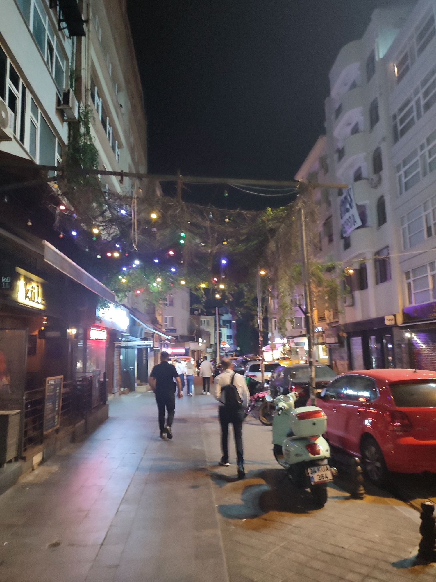 The image depicts a street scene at night in Kadıköy, Turkey, likely a commercial area. The perspective is from a pedestrian's viewpoint, looking down a sidewalk lined with buildings on the left and right. Decorative string lights are visible overhead, casting colorful illumination.
On the left side, there are storefronts, one with a prominent illuminated sign reading "kili". The architecture of the buildings suggests a mix of older and newer structures.
On the right side, there are multi-story buildings with balconies and windows. Several vehicles are present, including a red car parked along the curb and a light green scooter with a license plate that appears to read "34 KUA 364". Other vehicles, possibly cars and a motorcycle, are parked or moving further down the street.
Several individuals are walking on the sidewalk. Two men are prominently featured in the foreground and mid-ground. One man in a black t-shirt and dark pants is walking away from the camera. Another man in a white shirt and dark pants, carrying a backpack, is also walking away from the camera. Other pedestrians are visible in the background, suggesting moderate foot traffic. The ground is paved with stone tiles. The overall atmosphere is that of a lively urban street at night.