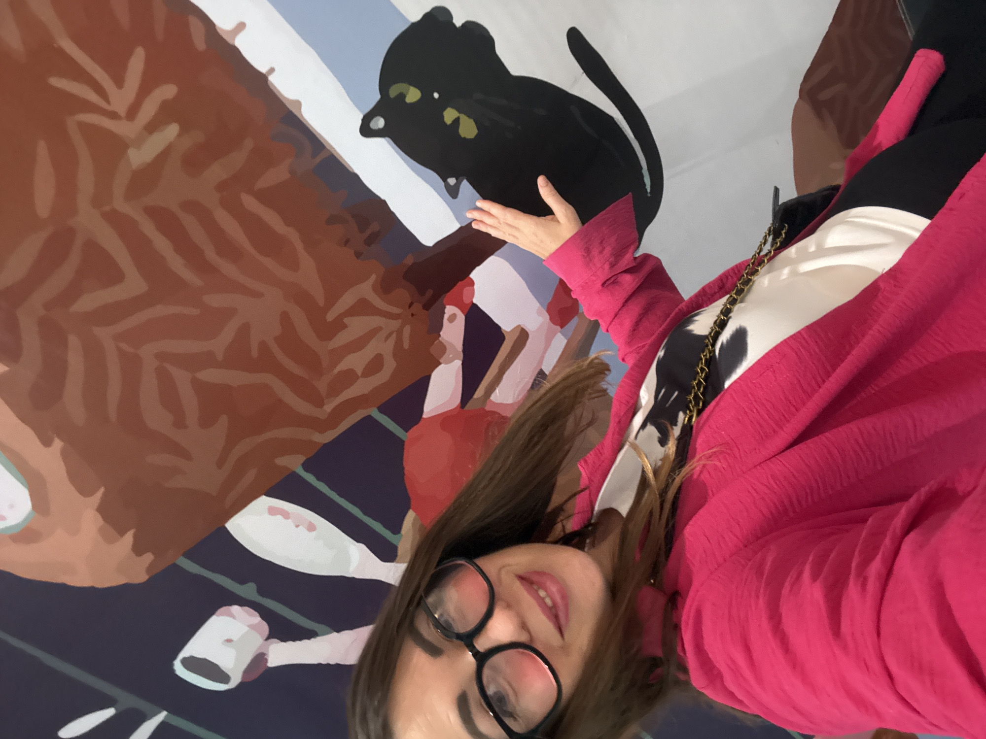 The image shows a woman in a bright pink blazer and black-rimmed glasses posing in front of a mural. She has long brown hair and is smiling at the camera. Her left hand is raised and touching the mural. The mural features a stylized depiction of a black cat with green eyes, and other abstract elements in brown, blue, and white. The scene appears to be indoors, possibly in a museum or gallery in Huizhou, China.