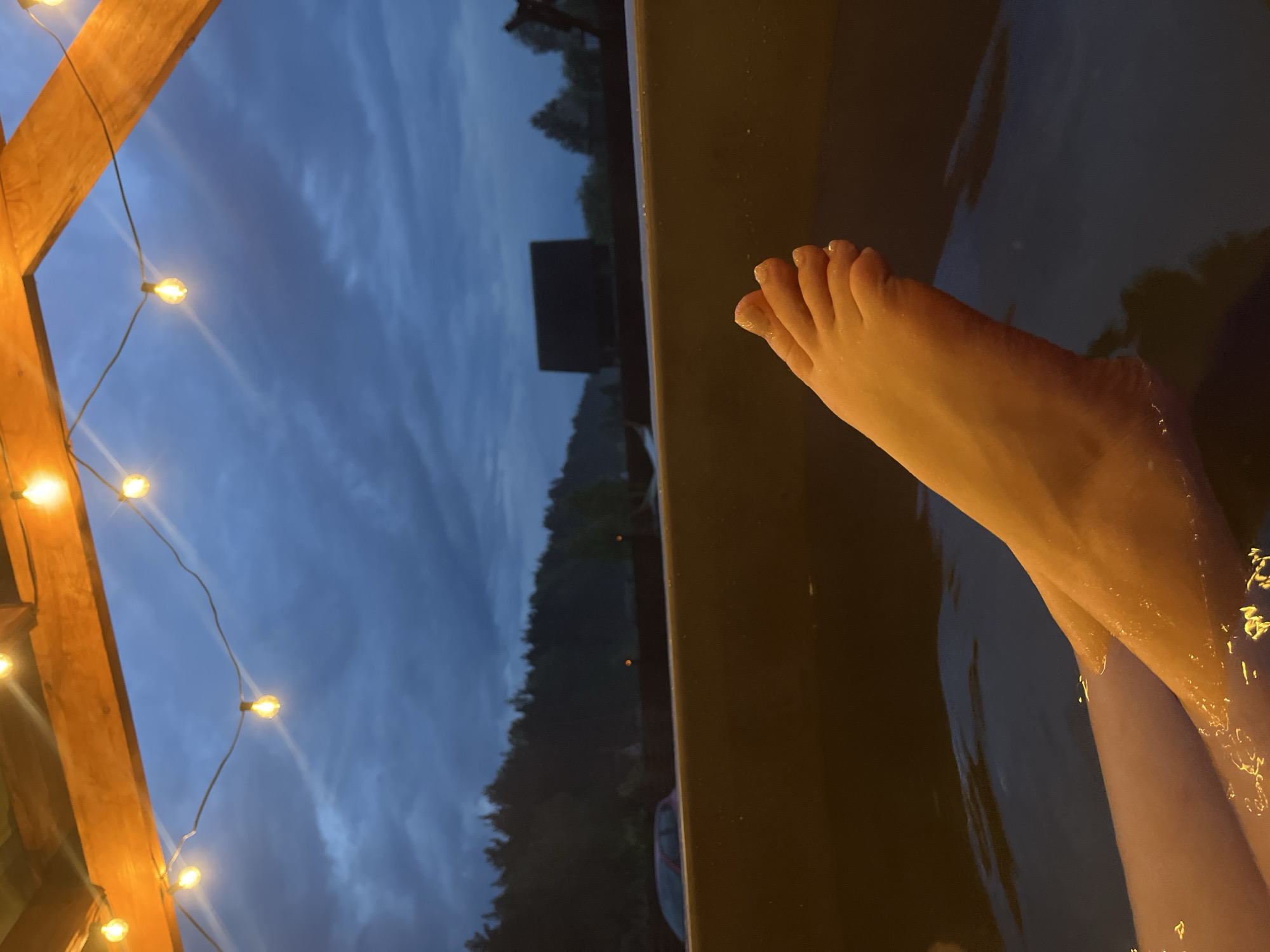 Here's a detailed description of the image:
A medium shot, rotated counter-clockwise, shows a foot and lower leg partially submerged in dark water, likely a hot tub or bath. The foot is positioned towards the upper right, with the toes pointing upwards. The skin appears slightly wet and reflective.
The edge of the hot tub, a dark, solid material, runs diagonally across the image, separating the water from the background. Beyond the edge is a blurred outdoor scene at dusk. Tall, dark trees line the horizon under a cloudy, blue-grey sky. A structure resembling a small building is visible among the trees.
To the left of the frame is a wooden structure with string lights hanging from it. The warm glow of the lights contrasts with the cool tones of the sky.
Given the availability of only an obscure location ("Гудагайскі сельскі Савет, Belarus"), it is impossible to independently verify if this is the location or if it is even in Belarus.