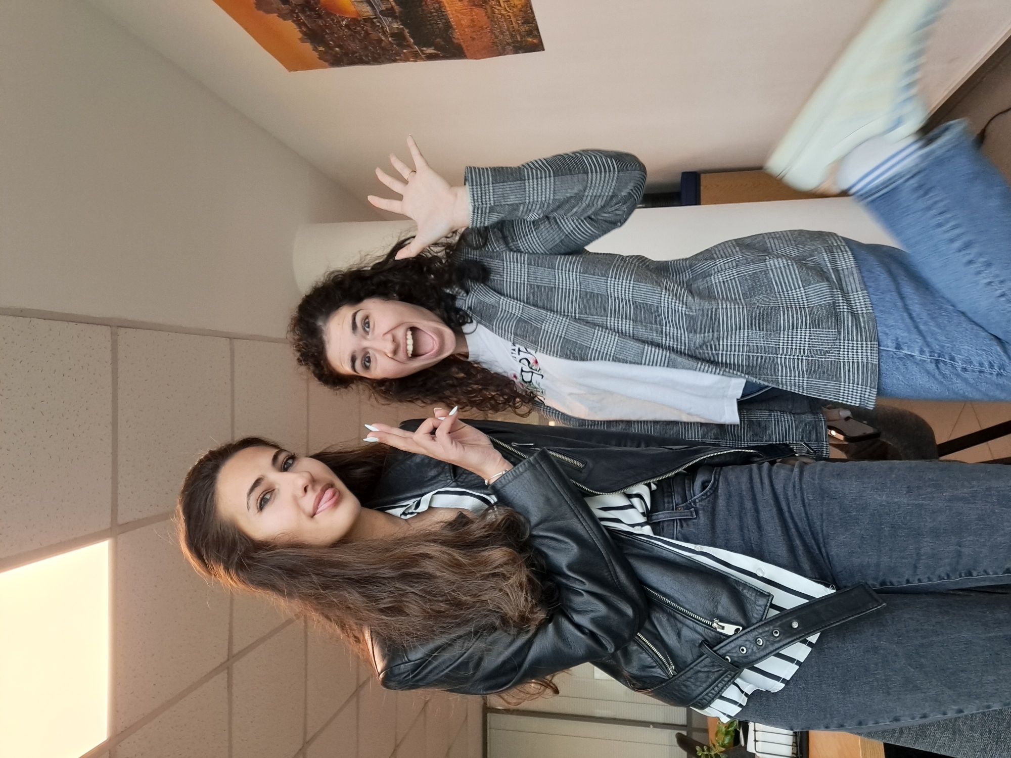 Two young women pose playfully for a photo indoors. The image is oriented vertically (originally taken in landscape, rotated 90 degrees clockwise).
In the foreground and to the left, a woman with long, dark brown wavy hair smiles at the camera, her tongue slightly extended. She wears a black leather jacket over a white and black striped shirt and dark gray jeans. Her right hand is raised, making a peace sign, and her fingernails are painted white.
Behind her and to the right, a second woman with dark curly hair appears to be laughing or shouting, her mouth wide open. She has both hands raised with fingers splayed in a playful gesture. She wears a gray plaid blazer over a white t-shirt with partially visible text that reads "ITALY," and blue jeans. Her left leg is raised, and she is wearing light-colored sneakers with blue stripes.
The setting appears to be a brightly lit room with white walls and an acoustic tile ceiling. A large, framed picture or poster depicting a city skyline, possibly at sunset or sunrise, hangs on the wall in the upper background. Bright natural light illuminates the scene, suggesting daytime. Both women exhibit happy and energetic emotions.