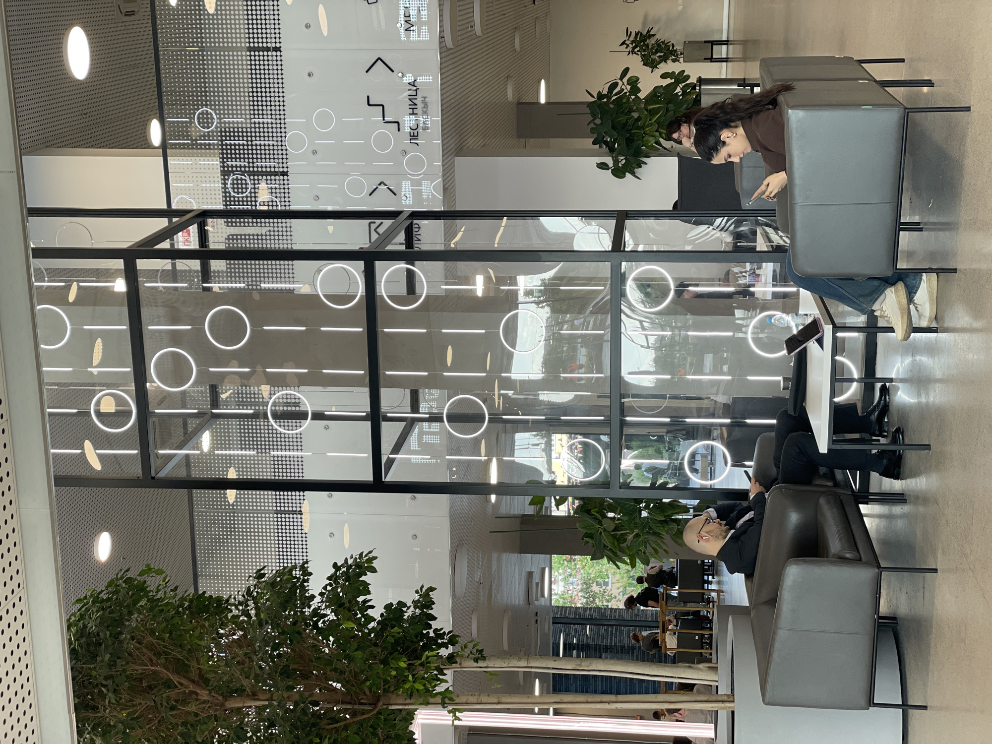 The image depicts the interior of a modern building, likely a business center or co-working space. The architecture features a prominent glass structure with illuminated circular and linear elements, creating a striking visual effect. In the foreground, two individuals are seated in plush, grey armchairs. On the left, a man in a dark suit is looking down at his phone, appearing contemplative. On the right, a woman with dark, curly hair is also engrossed in her phone. A low, white coffee table is positioned between them, holding a tablet and a water bottle.
The background reveals more of the building's interior, including other people seated at tables and a large, potted tree. The overall aesthetic is sleek and contemporary, with a focus on natural light and innovative design. The signage visible in the background, with the word "ЛЕСНИЦА" (which translates to "STAIRCASE" in Russian) and directional arrows, suggests the location is a public building. While the image does not explicitly state its location, the Russian language signage strongly indicates it is within Russia. Based on the architectural style and the presence of Russian text, this scene could plausibly be located in a modern building within the city of Kazan, Russia, perhaps a business center or a tech hub.