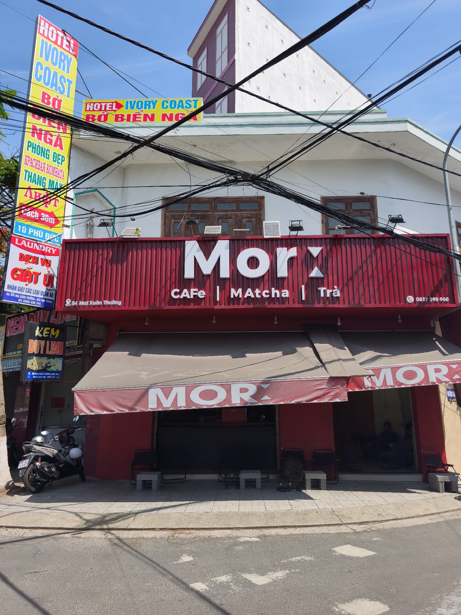 This is a medium shot of a storefront in the Bac Nha Trang Ward, Vietnam, on a sunny day. The building has a white facade with a bright red awning and a red sign above it that reads "Morx CAFE | MATCHA | Trà". The Morx sign features large white lettering and a phone number "0837 298 960" in the bottom right corner. A smaller red awning extends over the entrance, also with "MORX" in large white lettering.
To the left of the main storefront, a tall yellow and blue sign advertises "HOTEL IVORY COAST BO BIEN NGA PHONG DEP KRACHABAH KOMHATA THANG MAY AH PT". Below this, a red sign reads "LAUNDRY DICH VU GIAT UI".
In front of the store, a light grey motorcycle is parked on the sidewalk. The sidewalk is paved with light gray tiles. The street in front of the building is asphalt with some white markings. The building is adorned with numerous black electrical wires crossing overhead. The building itself appears to be three stories high with white walls and a dark roof. The second and third floors have windows.