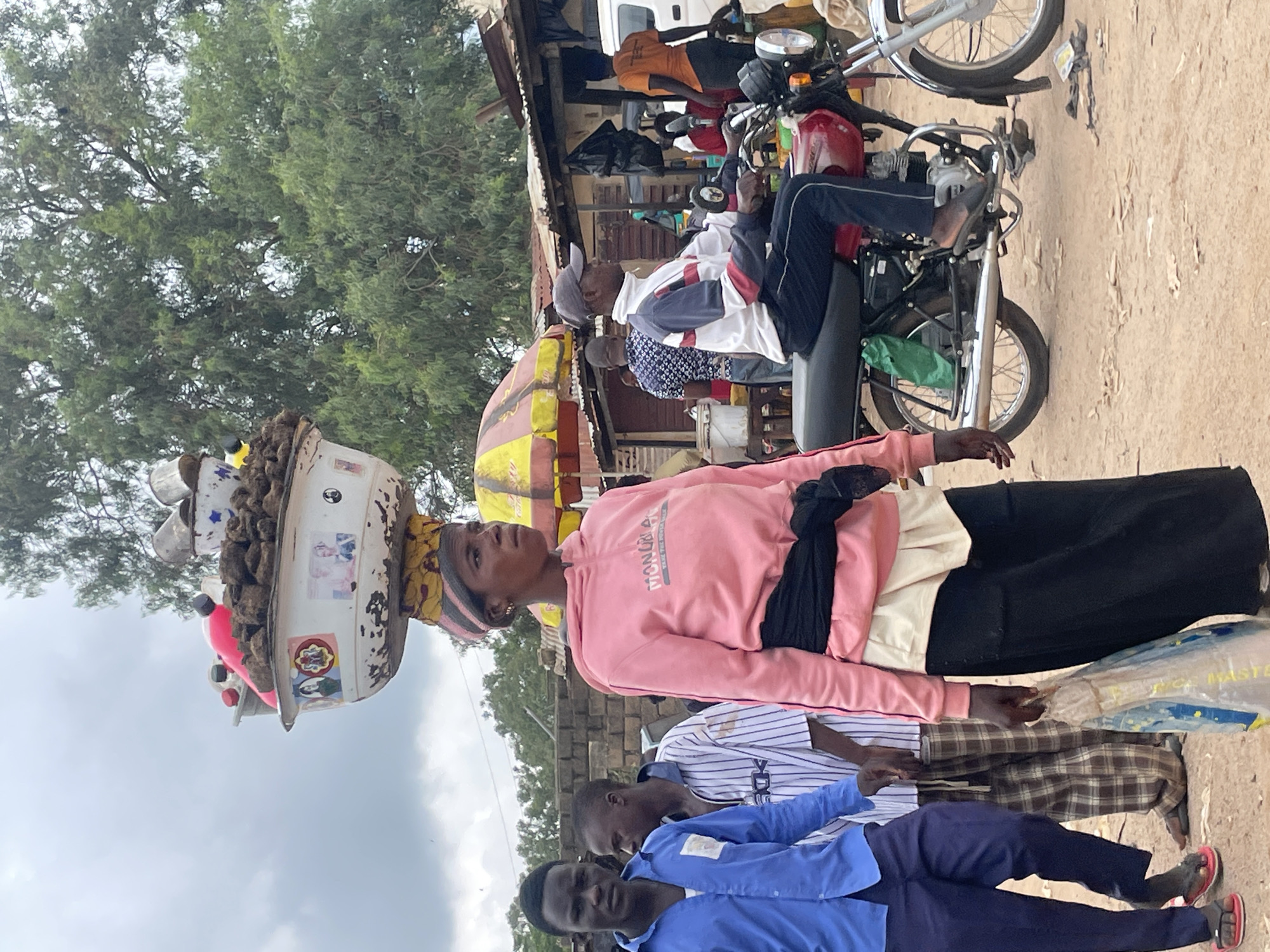 The image depicts a street scene in Wang, Nigeria, with a woman carrying a large bowl of goods on her head. She is wearing a pink hooded sweatshirt and a black belt. In front of her, a man in a blue shirt and dark pants walks with two other men. To the right, a man in a white, blue, and red jacket is sitting on a red motorcycle. Several market stalls and trees are visible in the background, suggesting a bustling environment. The ground is dusty, typical of a dry, outdoor market setting.