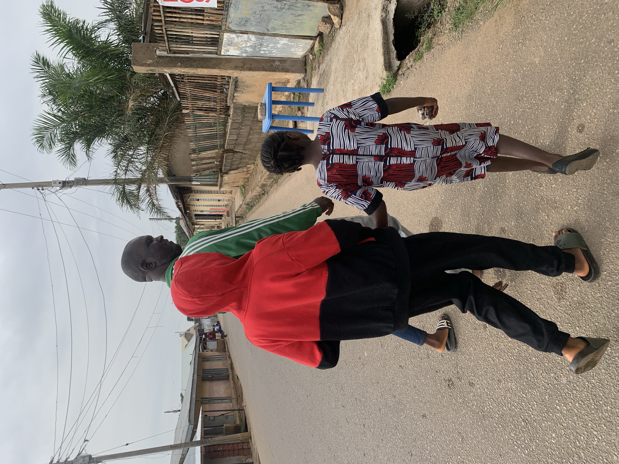 Three individuals walk away from the camera on a paved street in a Nigerian city, likely Jos. In the foreground, a child wearing a red, white, and black patterned dress is on the right, holding something in their right hand. To their left, a person in a red and black hooded sweatshirt and black pants walks ahead. To the left of the hooded figure, a man wearing a green and white striped jacket is partially visible. The street is lined with single-story buildings with corrugated metal roofs on the left and a fence with vertical wooden slats and some patchy grass on the right. Overhead, a network of electrical wires is strung between poles. A small blue table stands next to the fence on the right, with a brick on top. The sky is overcast.