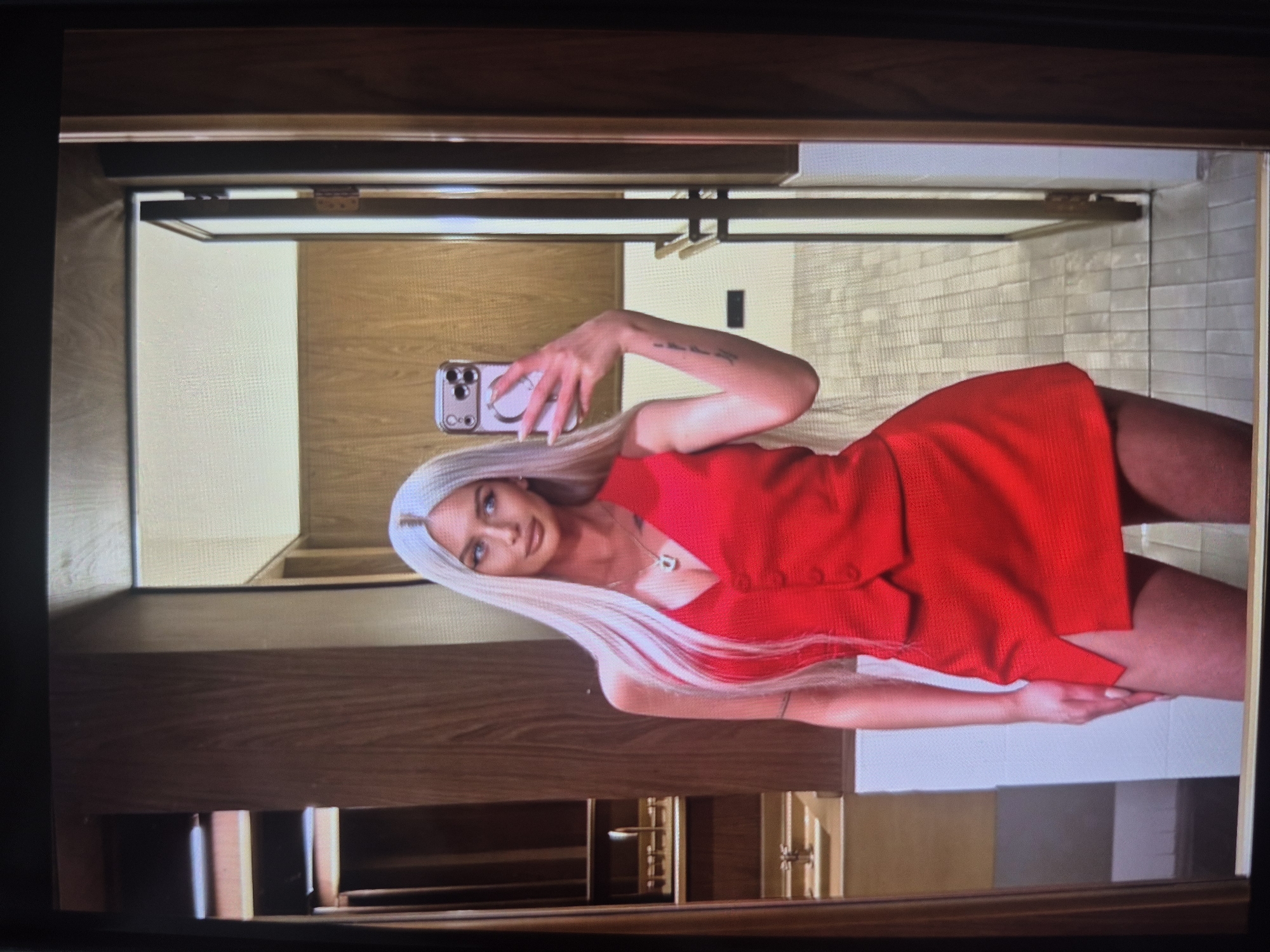 A woman with blonde hair and a red dress is taking a selfie in what appears to be a bathroom. The walls of the bathroom are tiled. The location is Tbilisi, Georgia.