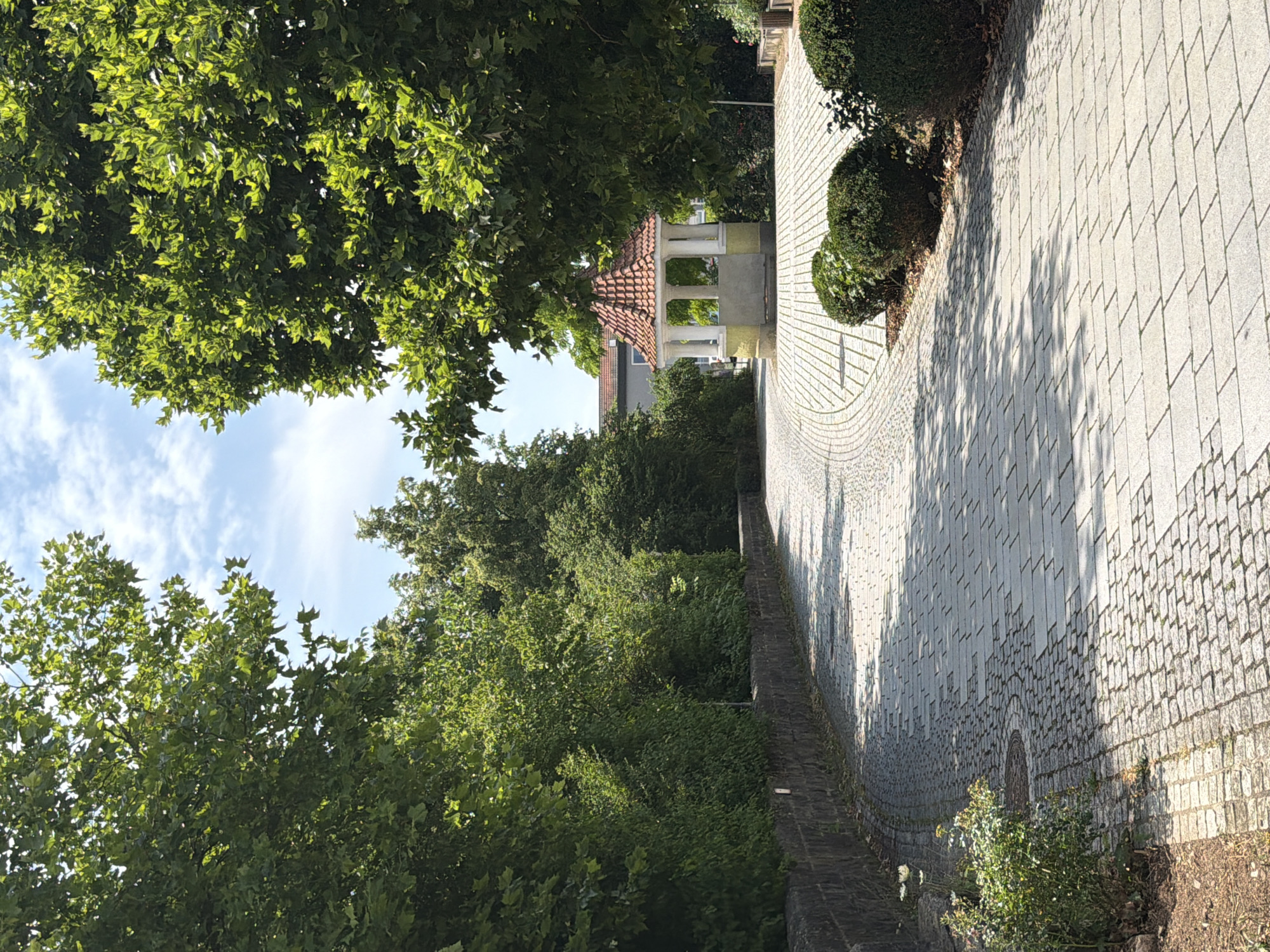 The image shows a paved pathway curving through a park-like setting. Large green trees line the pathway, casting shadows on the light-colored stone pavement. On the right side of the path, there are several rounded bushes. In the distance, a small, open-sided structure with a red tile roof supported by white columns is visible. The sky above is blue with some scattered white clouds. The pathway appears to be made of cobblestones and rectangular pavers. There are no people visible in the image. The scene suggests a public outdoor space in Ebermannstadt, Germany, likely a park or a garden area.