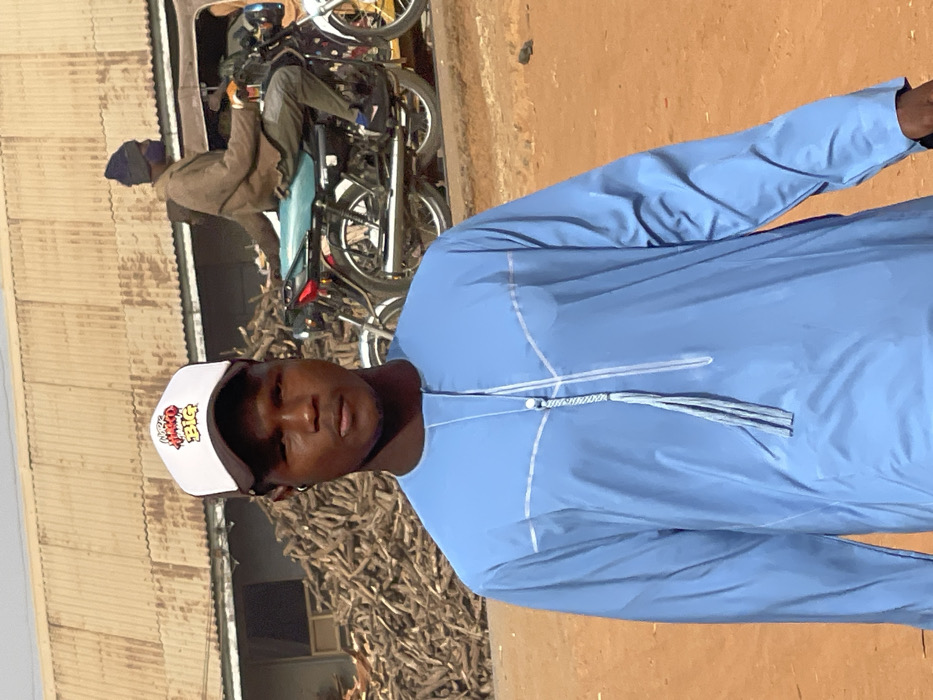 The image depicts an outdoor scene in Zaria, Nigeria. In the foreground, a young Black male is centered, facing the camera with a neutral expression. He wears a white and black baseball cap with multi-colored stylized text "AKO BIG" on the front, and a light blue long-sleeved traditional tunic featuring white piping details and a decorative tassel at the neckline. A small dark earpiece is visible in his right ear. The ground beneath him is reddish-brown dirt.
In the mid-ground, slightly to the left, is a large pile of light-colored dry wooden branches or firewood. To the right of this pile, a second Black male is seated on or operating a black motorcycle with a light blue seat, facing right, away from the camera. This individual wears a brown jacket, dark pants, a dark blue beanie, and a dark face covering. A second white motorcycle is partially visible behind the black one.
The background features a building with a light brown corrugated metal roof and walls, positioned behind the woodpile. The sky above is clear and light blue, indicating bright daylight conditions.