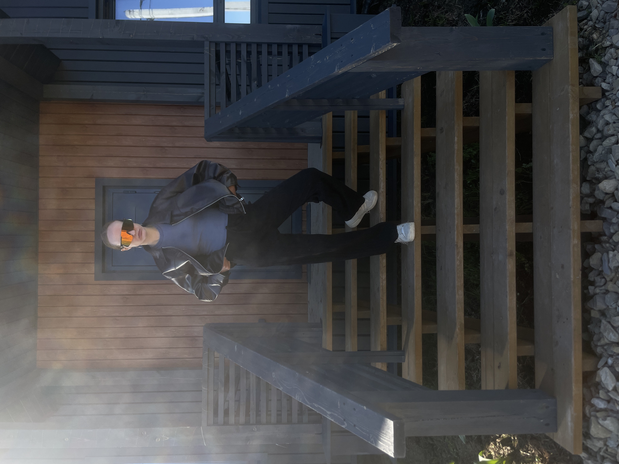 The image shows a person standing on a wooden staircase in Подшивалово, Russia. The staircase leads to a wooden building with a dark window frame. The person wears black pants, a blue t-shirt, a black leather jacket, white shoes, and orange sunglasses. The surrounding environment includes gravel and some vegetation.