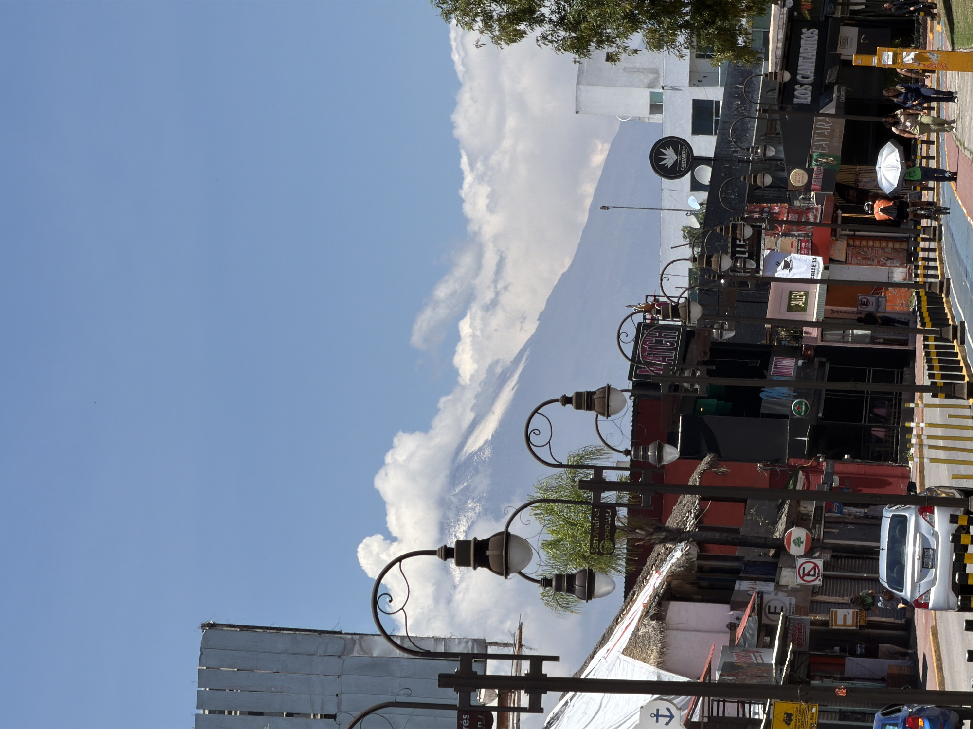 The image shows a street scene in San Andrés Cholula, Mexico, featuring a partial view of Popocatépetl volcano shrouded in clouds. The foreground includes streetlights, signage, buildings with thatched roofs, a variety of commercial storefronts, and pedestrians. A taxi is visible on the street.
