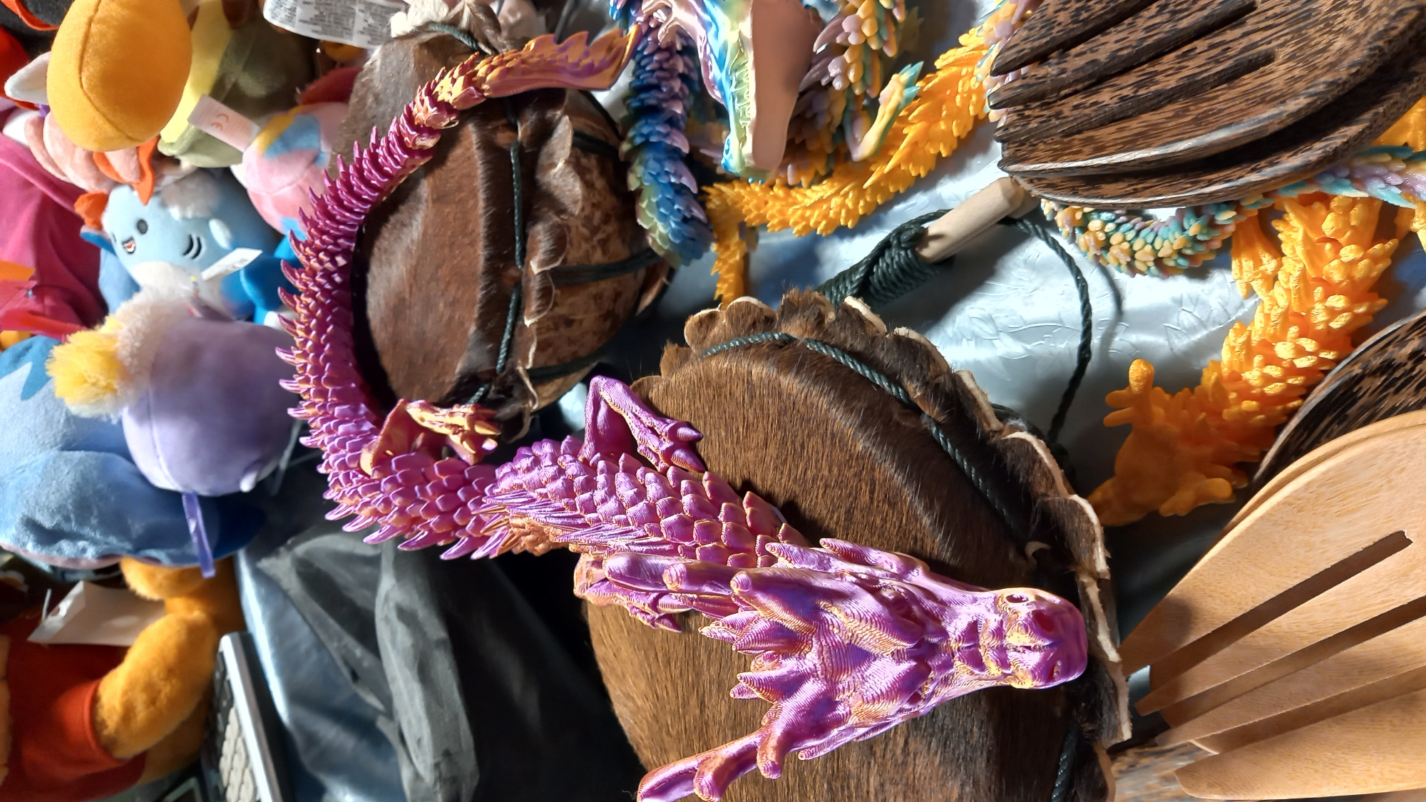 A purple and gold articulated dragon figurine rests on a brown, fur-covered drum. Another articulated dragon, in shades of blue, green, yellow, and purple, is partially visible in the background. A bright orange articulated dragon is also present. In the foreground, a wooden salad fork and tongs are laid out on a light blue, textured surface. Several stuffed animals, including Winnie the Pooh and a blue character with yellow accents, are scattered in the background. The scene appears to be an indoor setting.