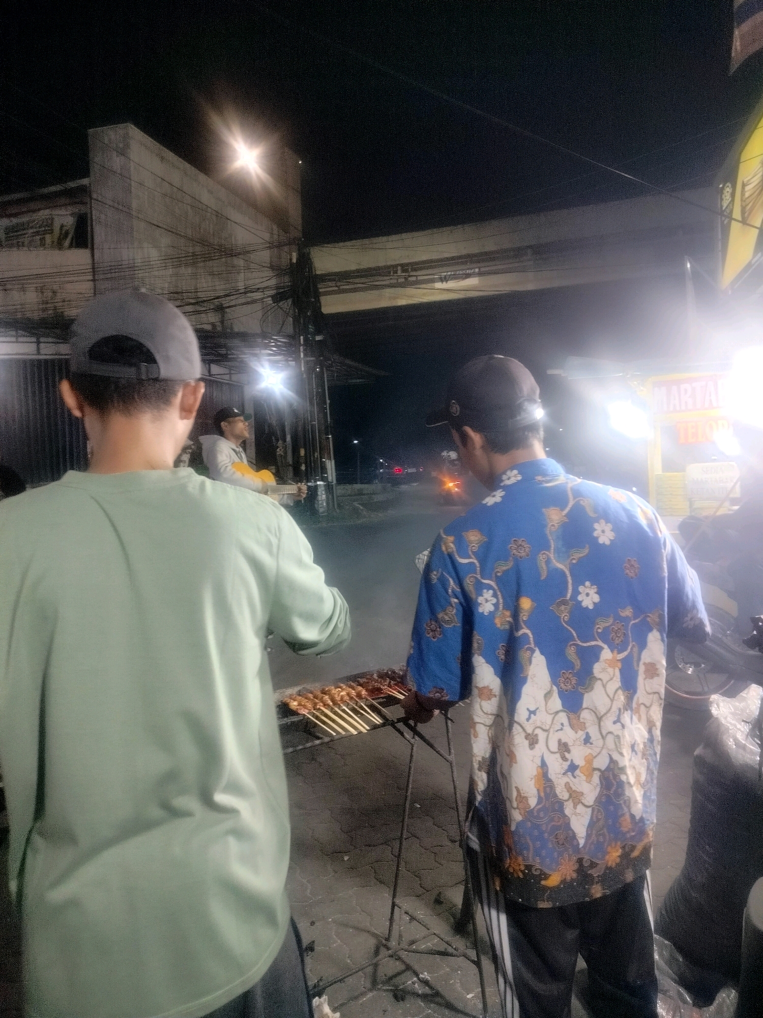 The image depicts an outdoor night scene in the city of Bekasi, Indonesia, likely near a roadside or market area. In the foreground, two individuals are visible from behind. The person on the left wears a light green long-sleeved shirt and a grey baseball cap. The person on the right wears a blue batik shirt with a white and orange floral pattern and a black baseball cap. Both are facing away from the camera, towards a grill where skewers of food are being cooked over charcoal. The background reveals a street scene with a building structure overhead, likely an overpass or elevated road, supported by pillars. Numerous electrical wires are strung across the scene. Further in the background, another person is seen holding a yellow object, possibly a guitar, and is illuminated by a bright light. A yellow sign with text in Indonesian, including "MARTABAK TELO..." is visible on the right, suggesting a food stall. The ground is paved with a rough, uneven surface. The overall atmosphere is of a lively, informal evening gathering or street food environment.
