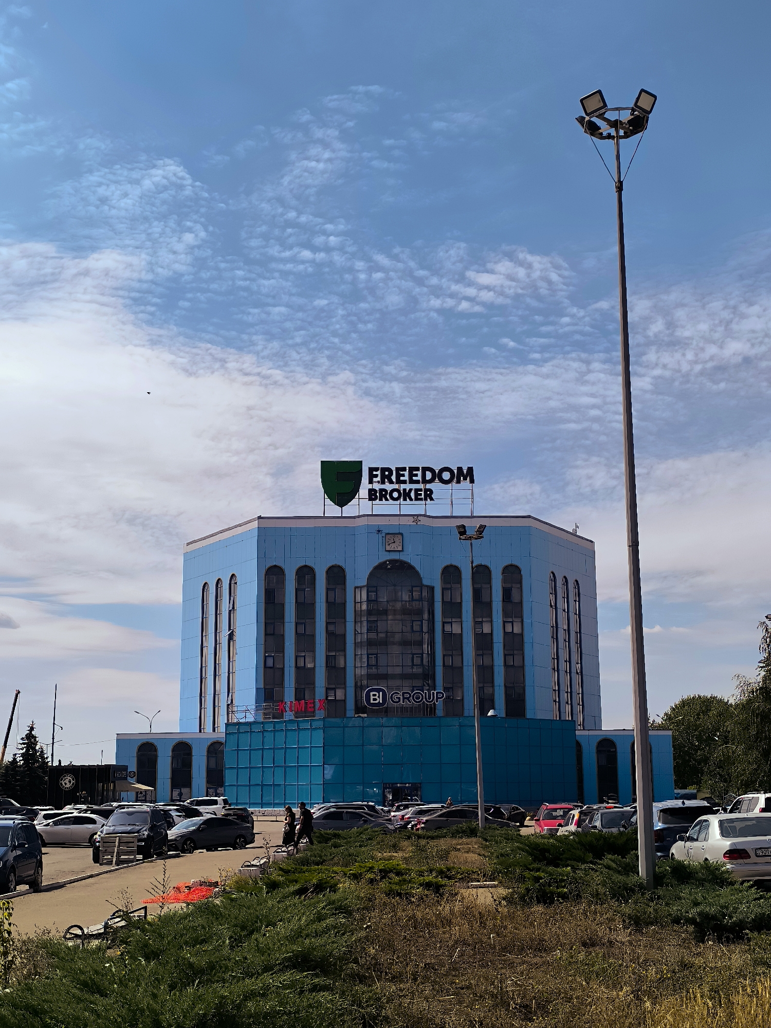 The image shows a bright, multi-story blue building with a hexagonal base and a rounded top, prominently displaying the signage "FREEDOM BROKER" with a green shield logo above it. The building has numerous windows and a section with a darker blue glass facade. Below the "FREEDOM BROKER" sign, the letters "BI GROUP" are visible on the darker blue section. The building is situated in a city with a parking lot in front, filled with various cars. The scene is captured on a clear day with a blue sky dotted with white, wispy clouds. A tall lamppost with multiple lights stands to the right of the building. In the foreground, there is dense green foliage and dry grass. The location is identified as the city administration building in Qarağandı, Kazakhstan.
